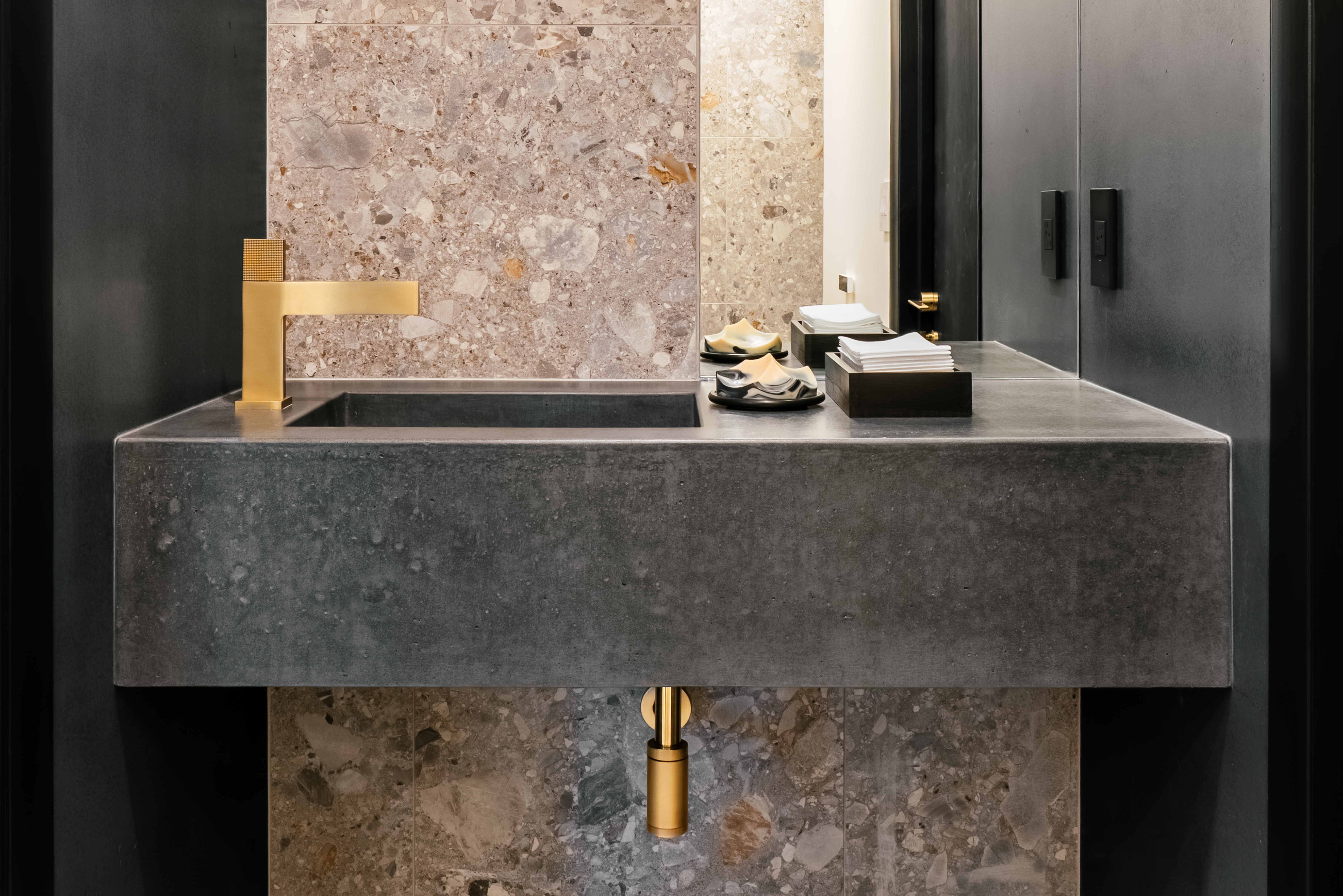 A modern bathroom vanity with a dark gray concrete integrated sink and countertop, a minimalist brass faucet, and a speckled terrazzo-style stone tile backsplash. Decorative soaps and folded towels sit on the counter beside a mirror, reflecting dark cabinetry.