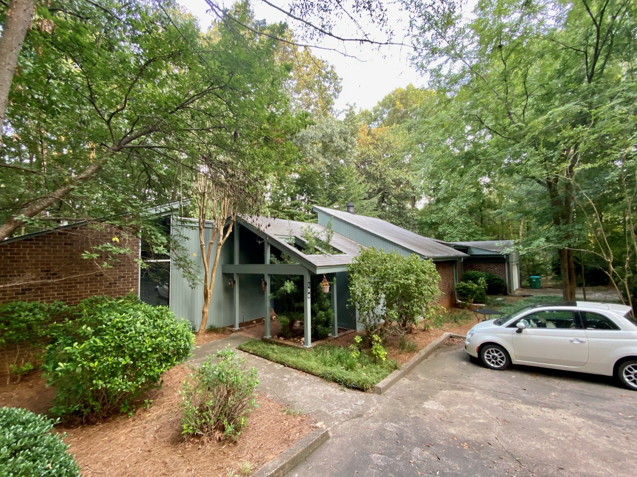 An exterior view of a mid-century modern home with muted green siding and prominent angled rooflines, surrounded by dense green trees and landscaping, with a white car parked in the driveway.
