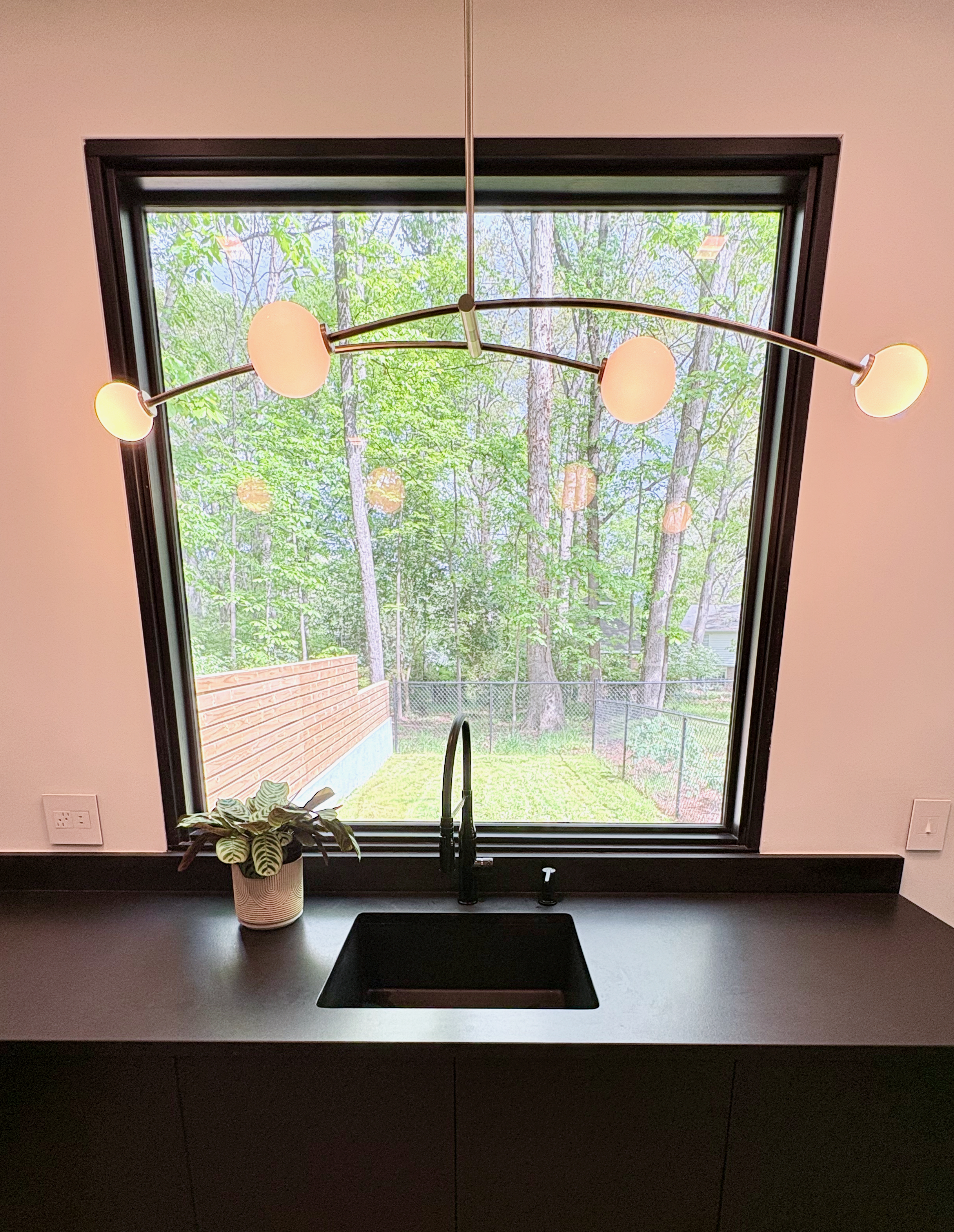 A modern kitchen sink area features a large, trapezoidal window looking out onto a lush green backyard with trees and a wooden fence. A contemporary chandelier with five spherical lights hangs above the dark countertop, which holds a black sink, a matte black faucet, and a potted plant.
