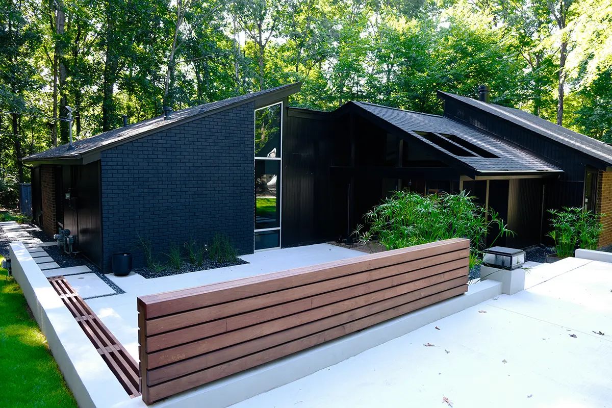 A modern black house, Ravenwood, with angular rooflines and large windows, is surrounded by lush green trees and features a contemporary concrete and wood-slat landscape design in Athens, GA.