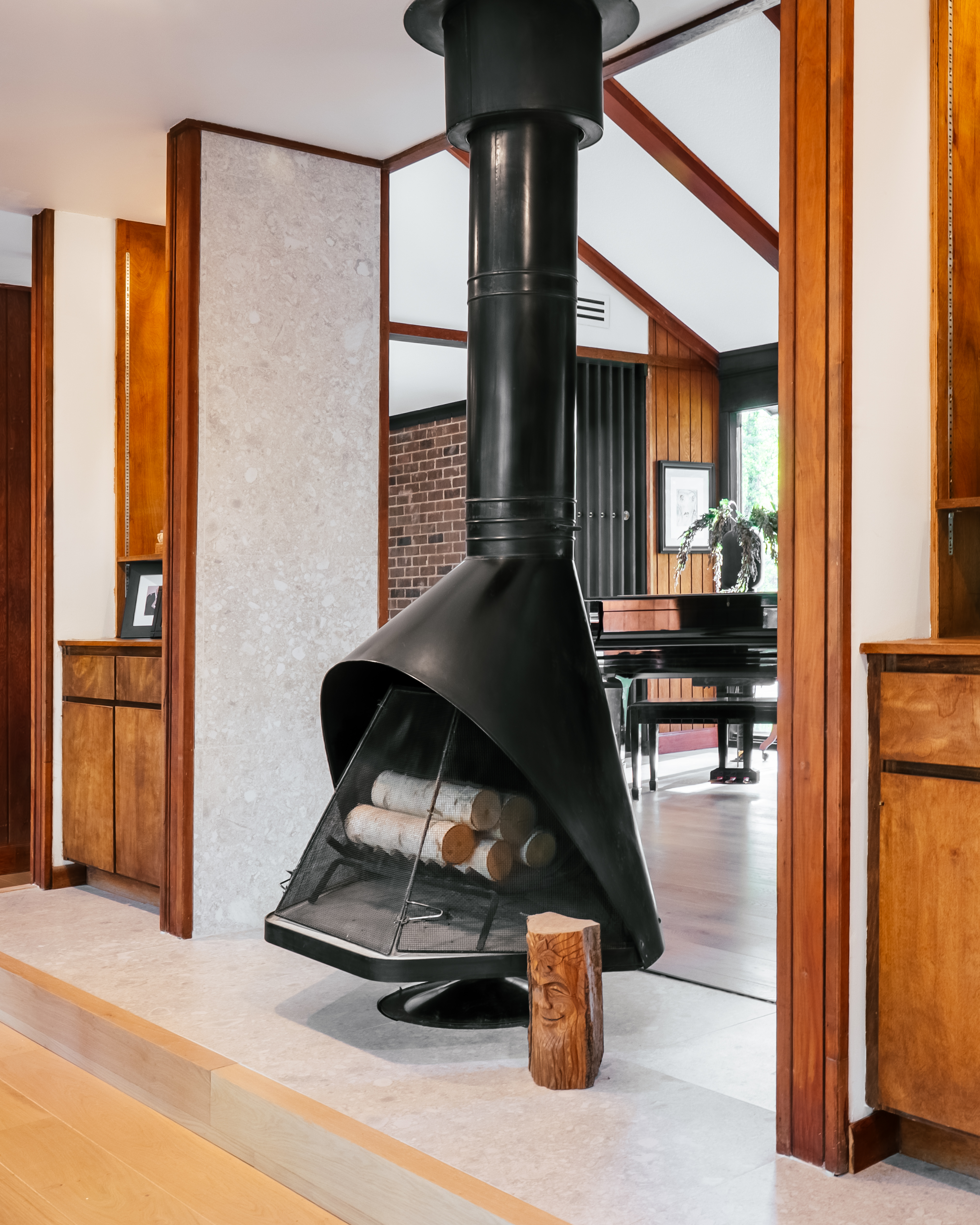A black suspended fireplace with birch logs sits on a light gray tile floor, framed by wooden door frames and a textured wall, with a grand piano visible in the background.