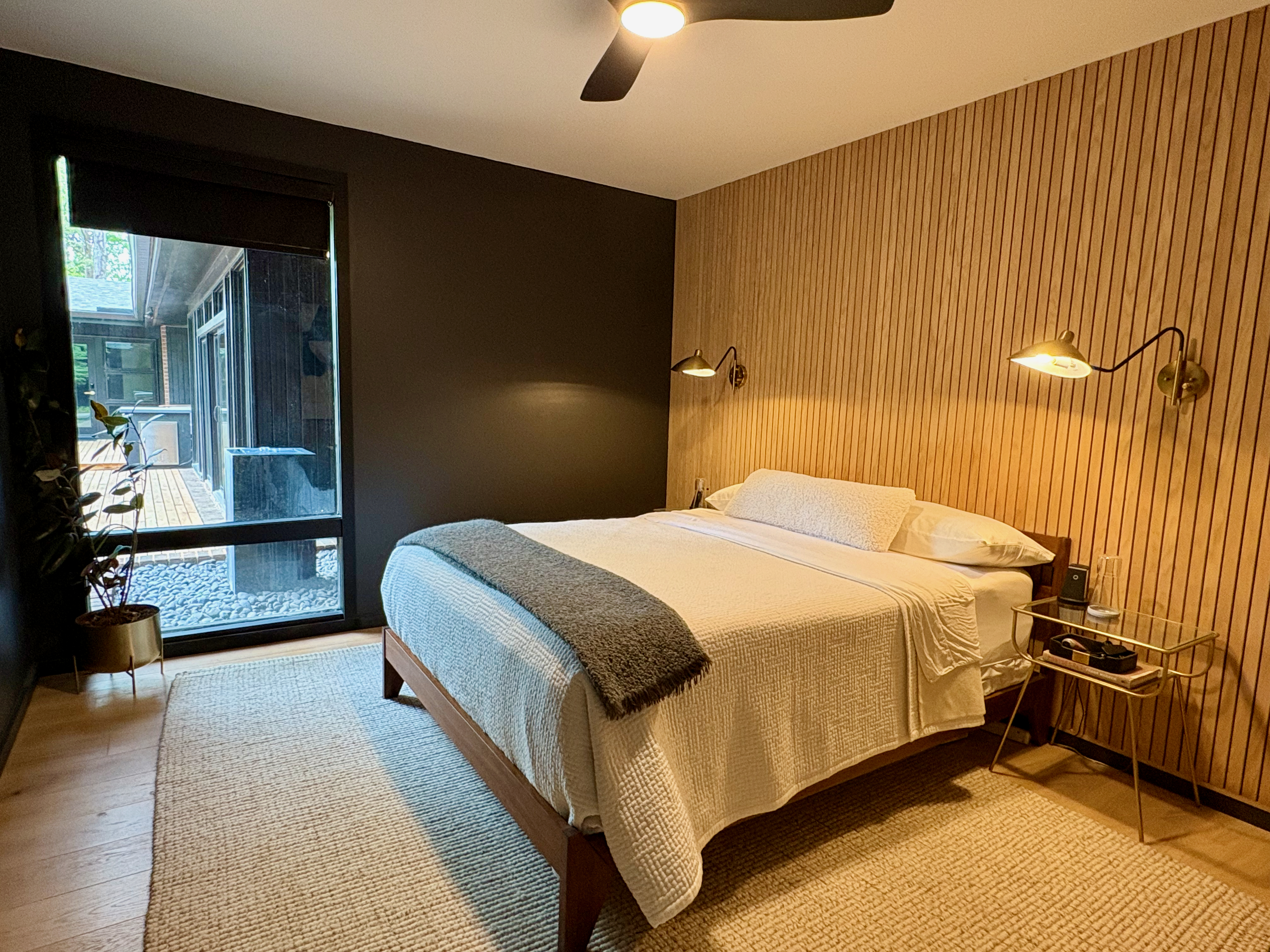 A luxurious mid-century modern bedroom featuring a large bed with white bedding, a dark charcoal accent wall, and a striking slatted wood panel wall behind the bed, illuminated by brass sconces. A large window offers a view of an outdoor deck and natural landscaping, enhancing the room's serene ambiance.