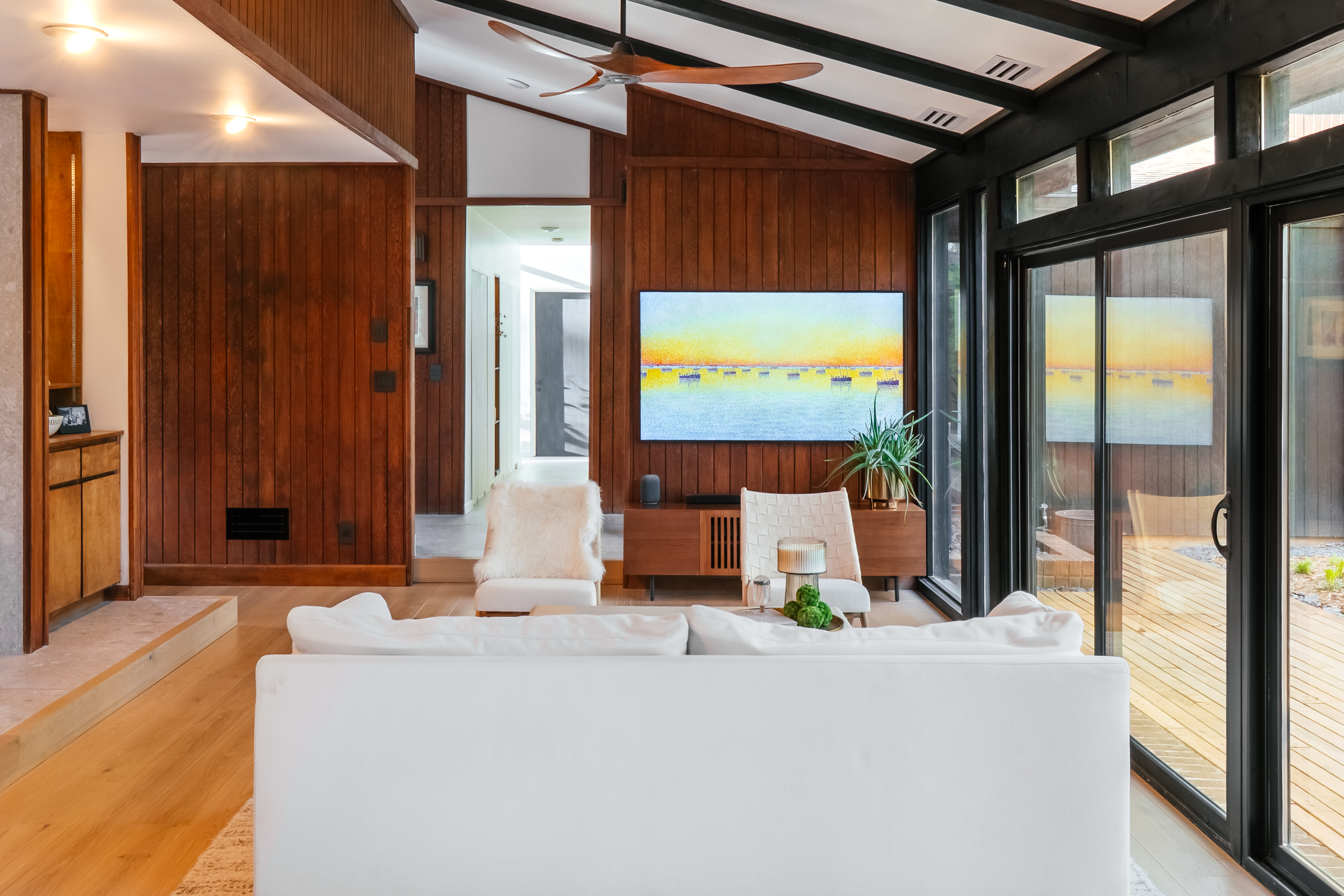 A luxurious mid-century modern living room featuring warm wood paneling, a vaulted ceiling with a wooden fan, a large white sofa, and a television displaying abstract art, with glass doors opening to an outdoor deck.
