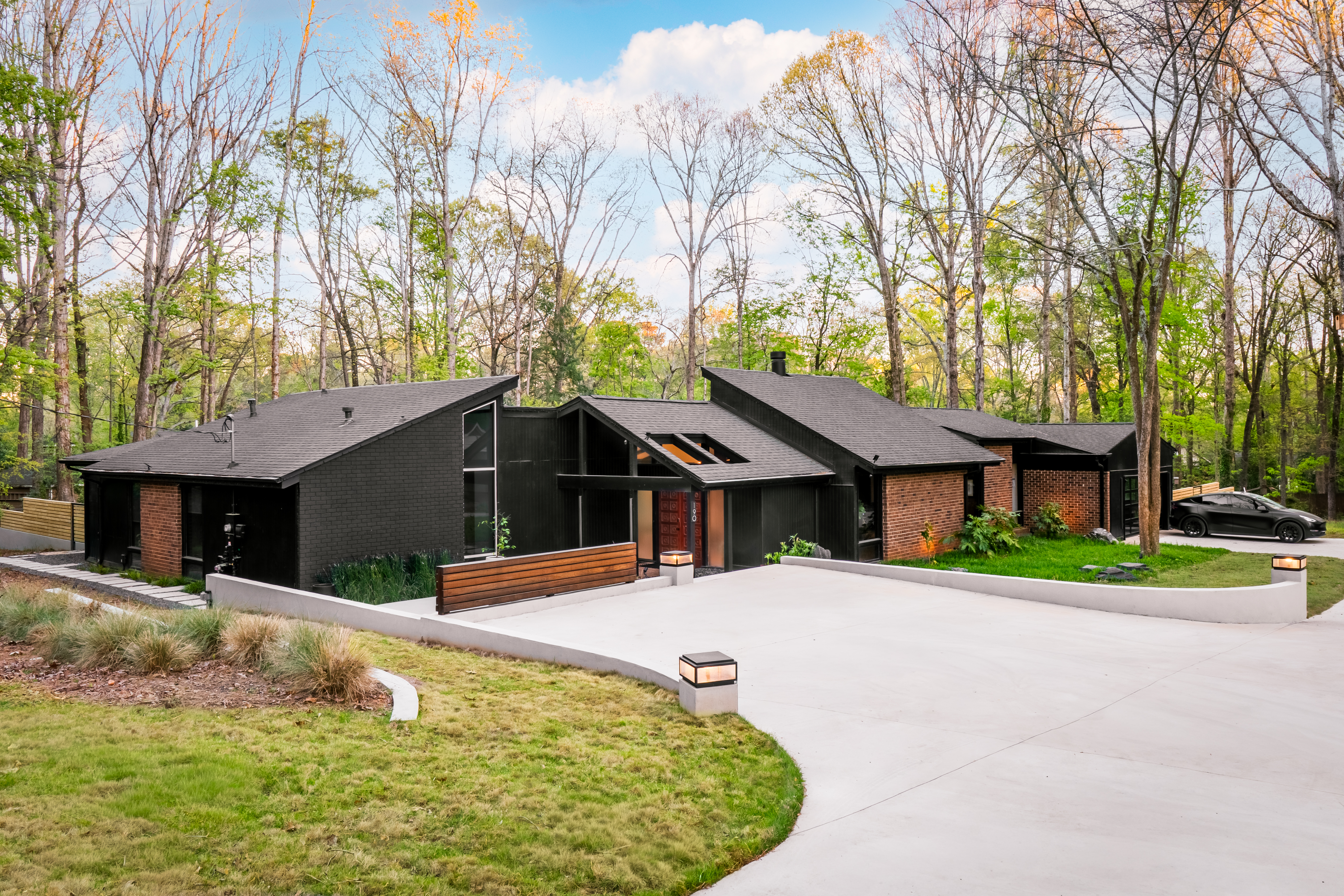 An exterior wide shot of Ravenwood, a dark-colored mid-century modern home with black brick and vertical wood siding, a complex angular roof, and large windows, situated amidst a wooded landscape with a wide concrete driveway leading to the entrance.