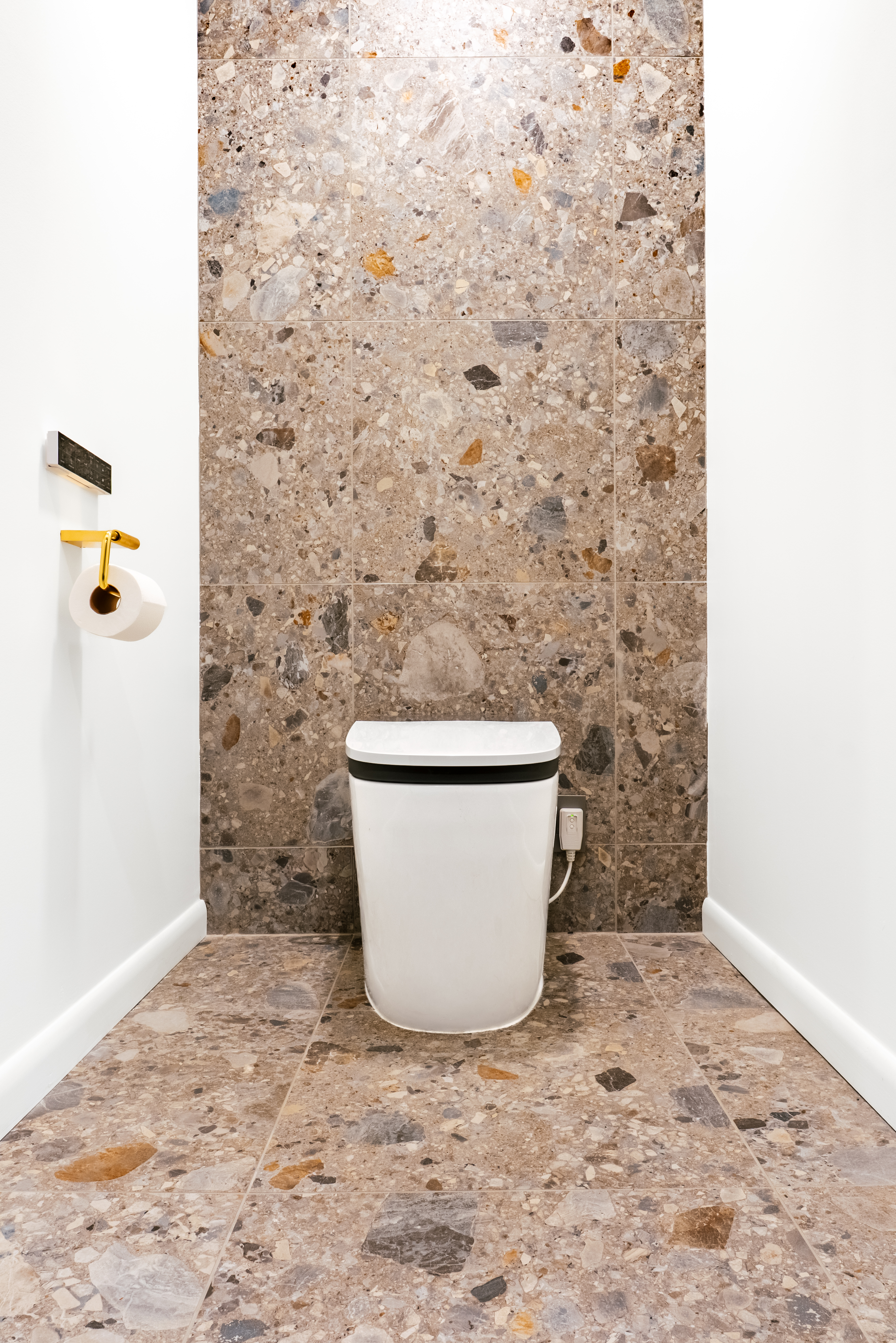 A minimalist powder room features a white, modern toilet set against a wall and floor covered in large, square terrazzo-style tiles with diverse stone fragments in shades of grey, brown, and ochre. A gold toilet paper holder is visible on the white wall to the left.