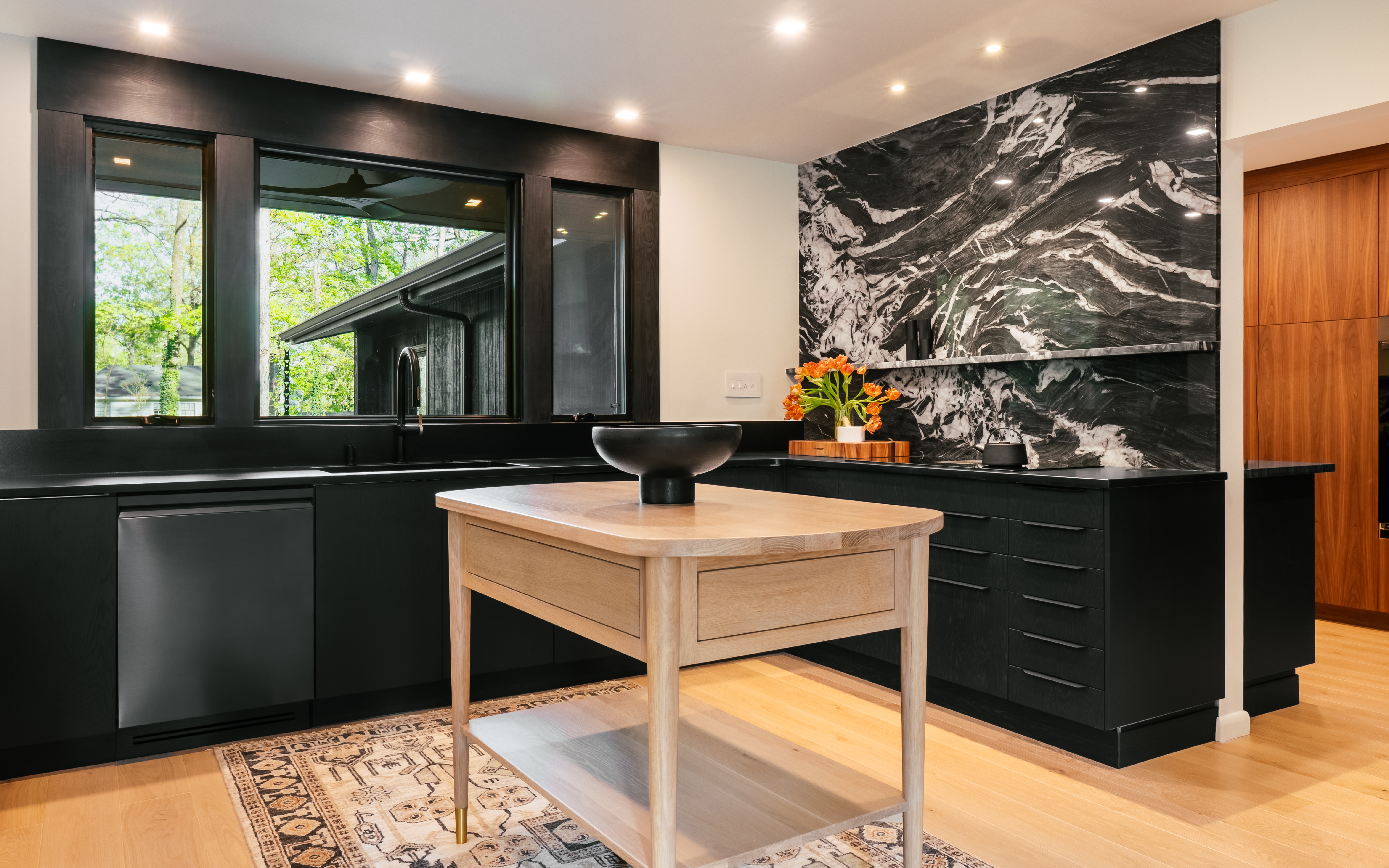 A modern kitchen featuring dark black cabinetry, a bold black and white marble backsplash, and a light wooden island, with large windows offering a lush green view of the outdoors.