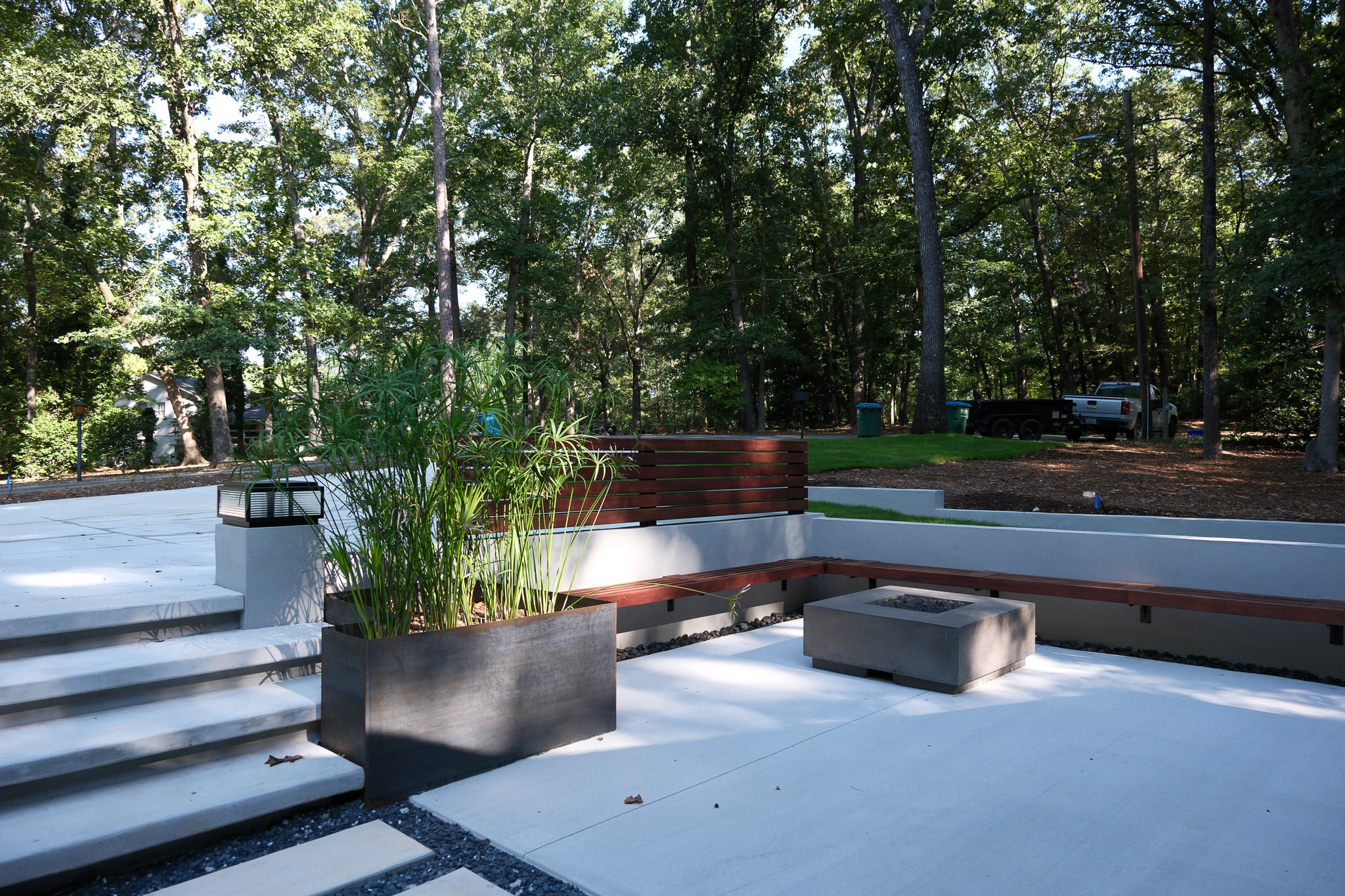 A modern outdoor patio with concrete steps, a large metal planter with tall green plants, a built-in wooden bench, and a rectangular concrete fire pit, all set against a backdrop of dense, mature trees.