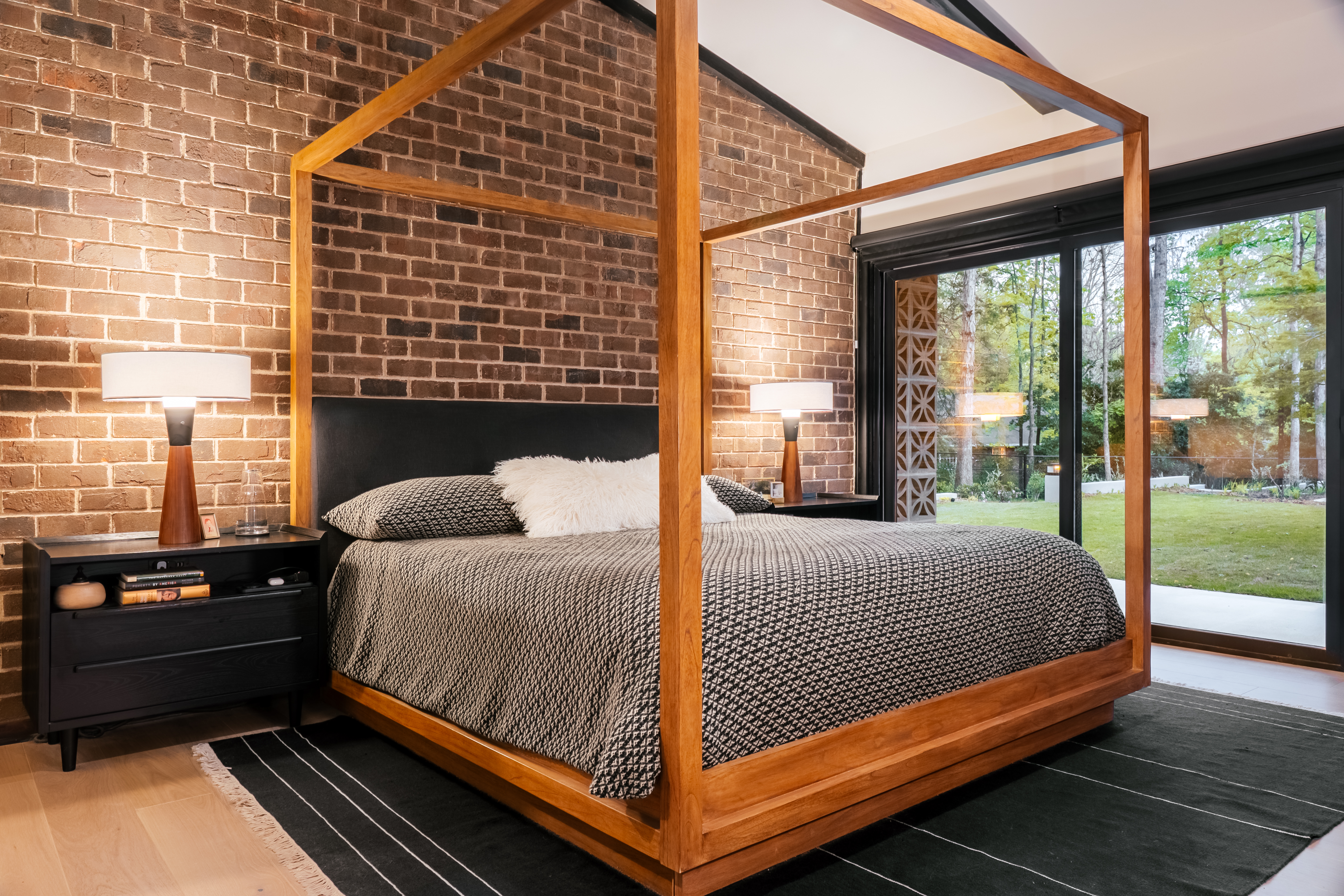 A luxurious bedroom featuring a prominent exposed brick wall, a modern wooden four-poster bed with black and white bedding, and large glass sliding doors opening to a lush green backyard with trees.