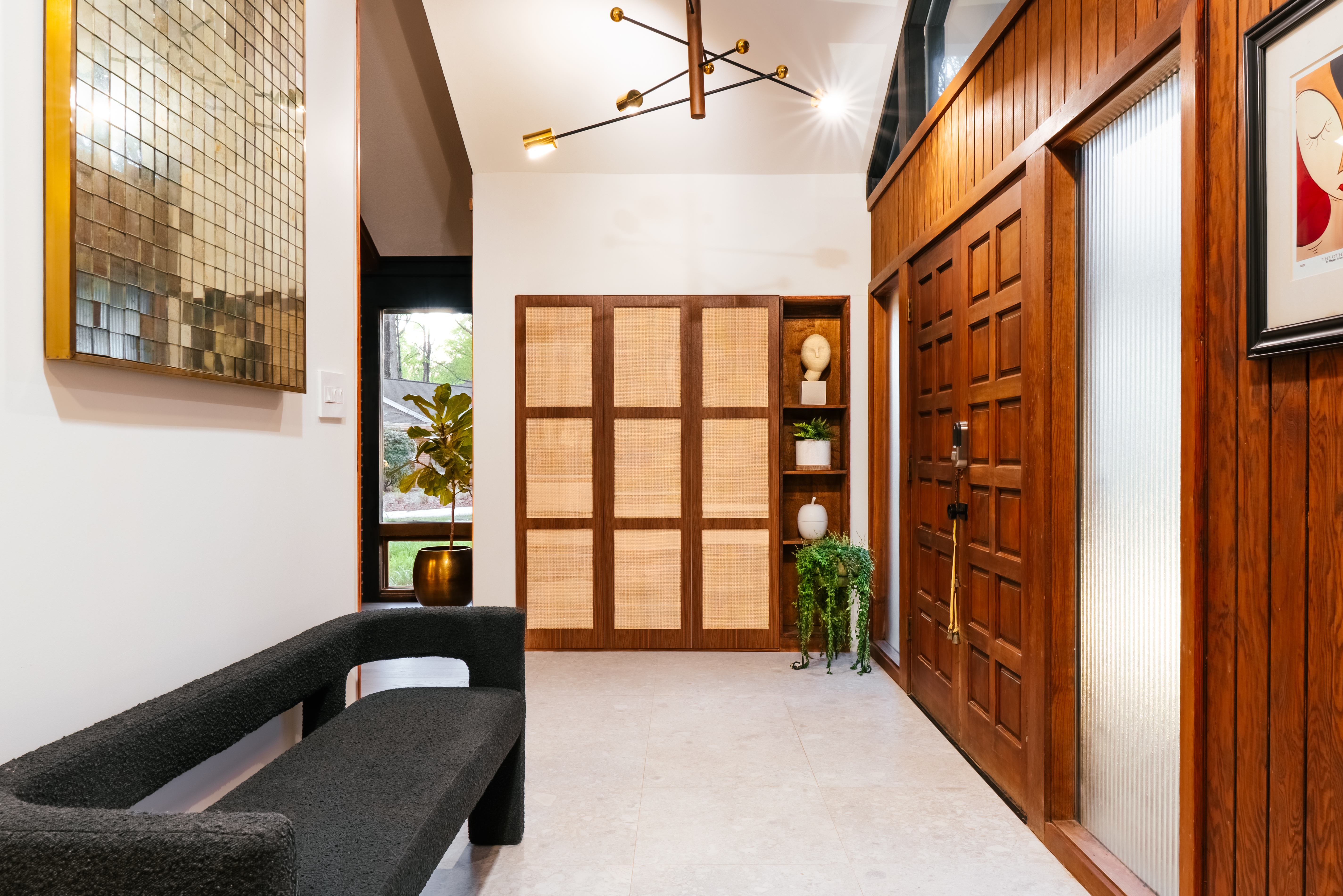 A stylish mid-century modern entryway features a dark wood paneled door, light gray tiled floor, a black textured bench, and cane-front built-in cabinets with decorative shelves, all illuminated by a unique sputnik-style chandelier.