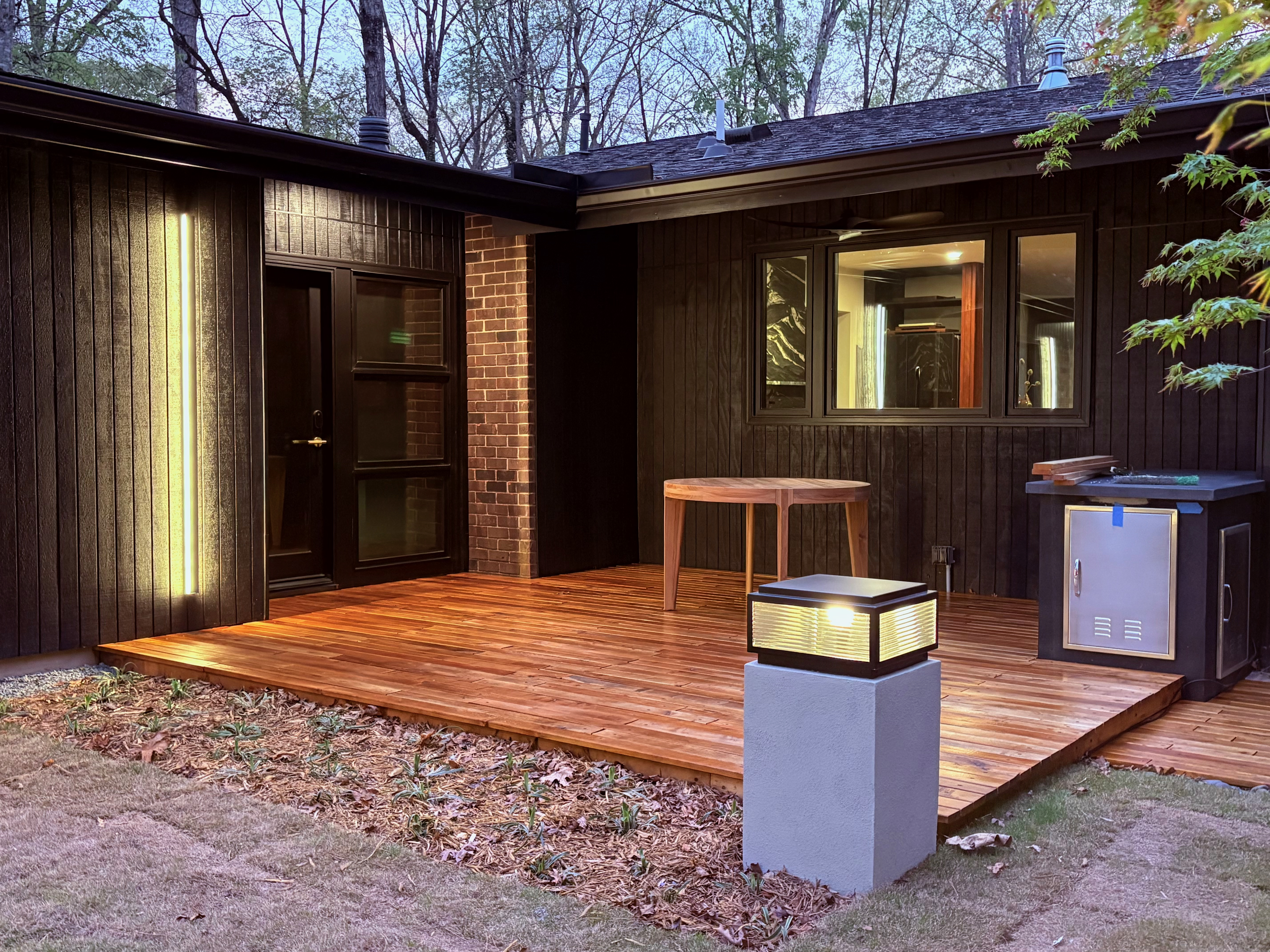 An evening view of Ravenwood's mid-century modern home in Athens, GA, showcasing a warm wooden deck with contemporary lighting, dark wood siding, a brick column, and an outdoor grill, nestled against a backdrop of trees.
