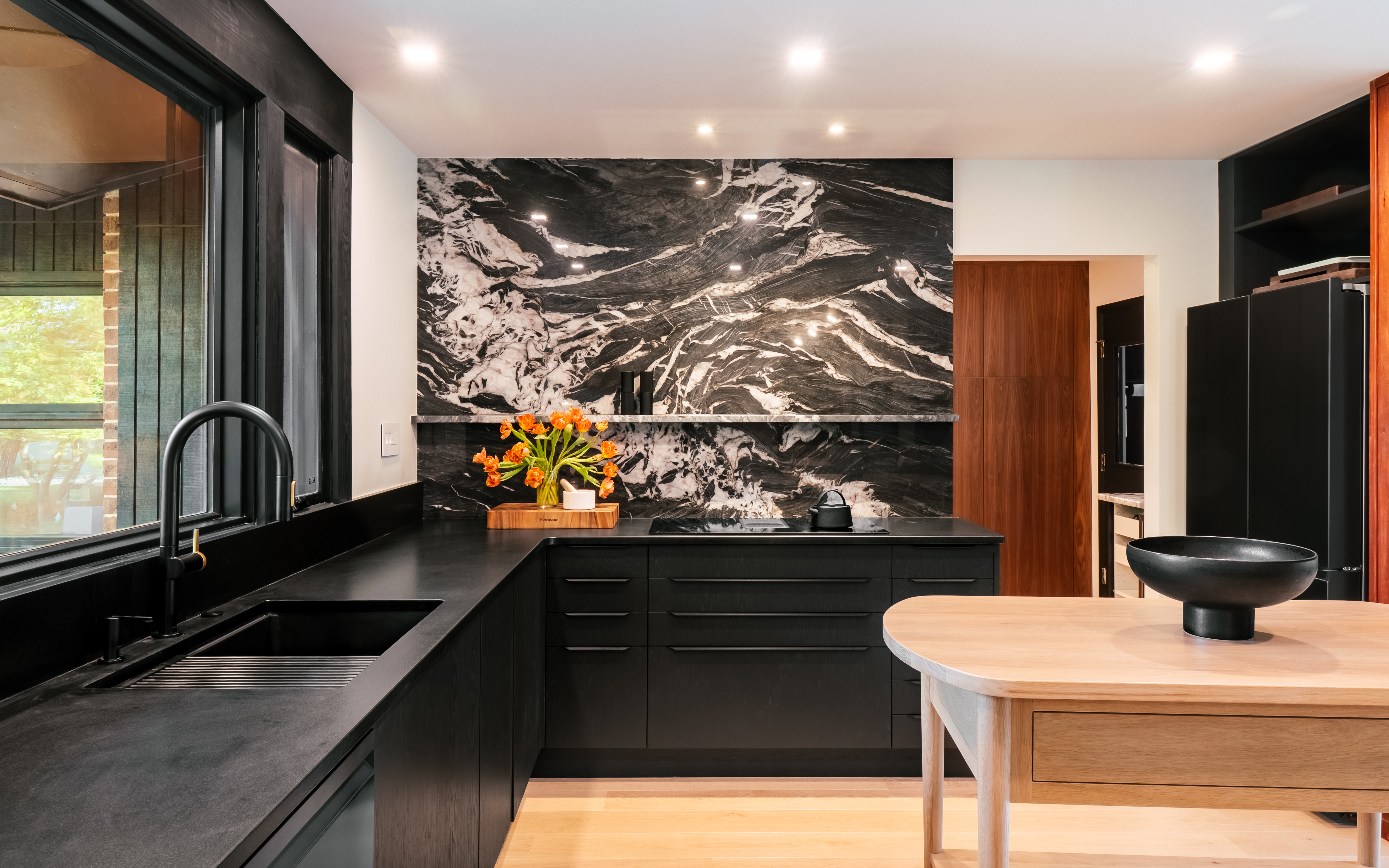 A modern kitchen featuring black cabinetry, countertops, and a striking black marble backsplash with white veining, illuminated by recessed lighting and natural light from a large window overlooking greenery.