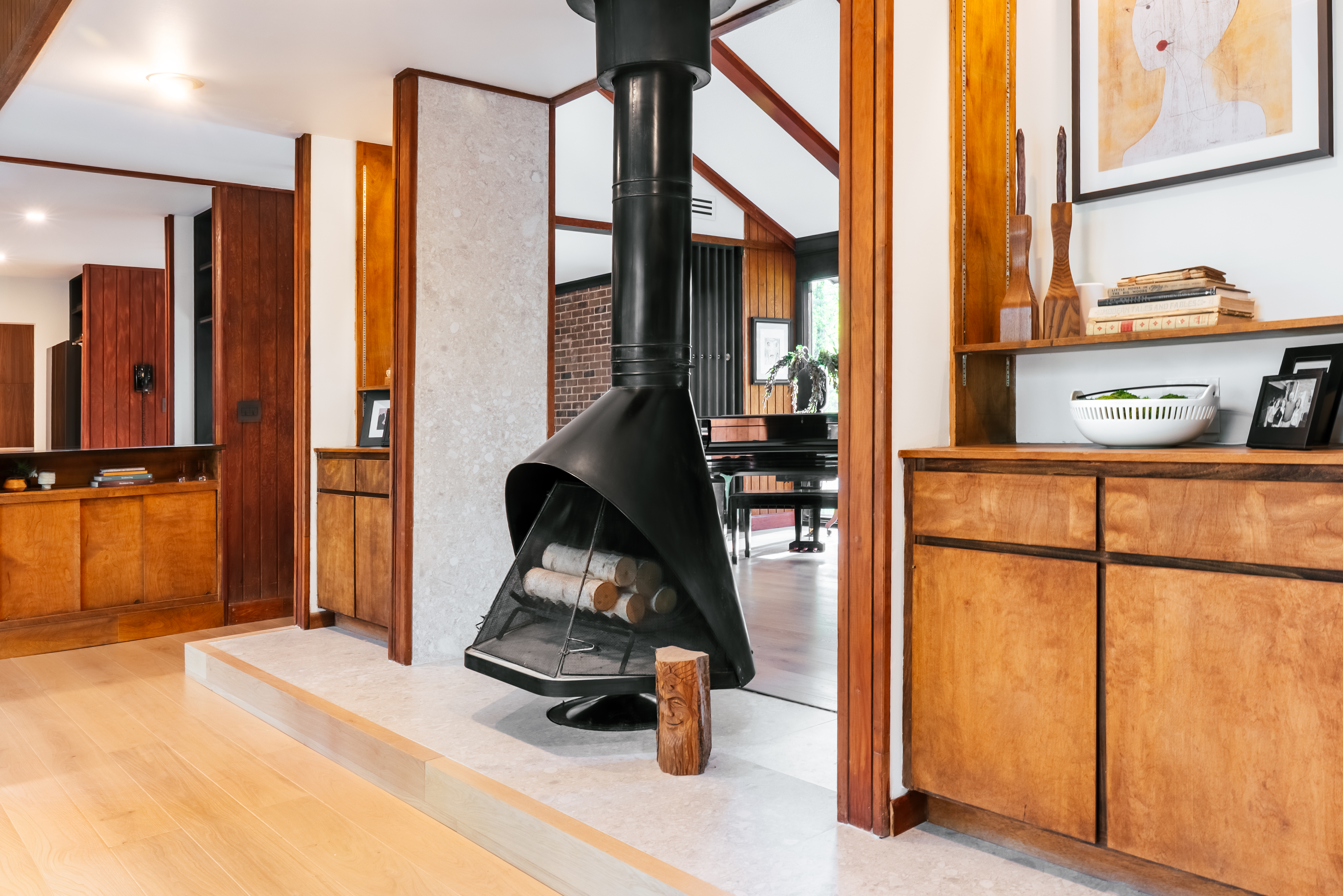 A stylish mid-century modern interior featuring warm wood paneling and cabinetry, light hardwood floors, and a striking black suspended fireplace with logs, leading into another room with a grand piano.