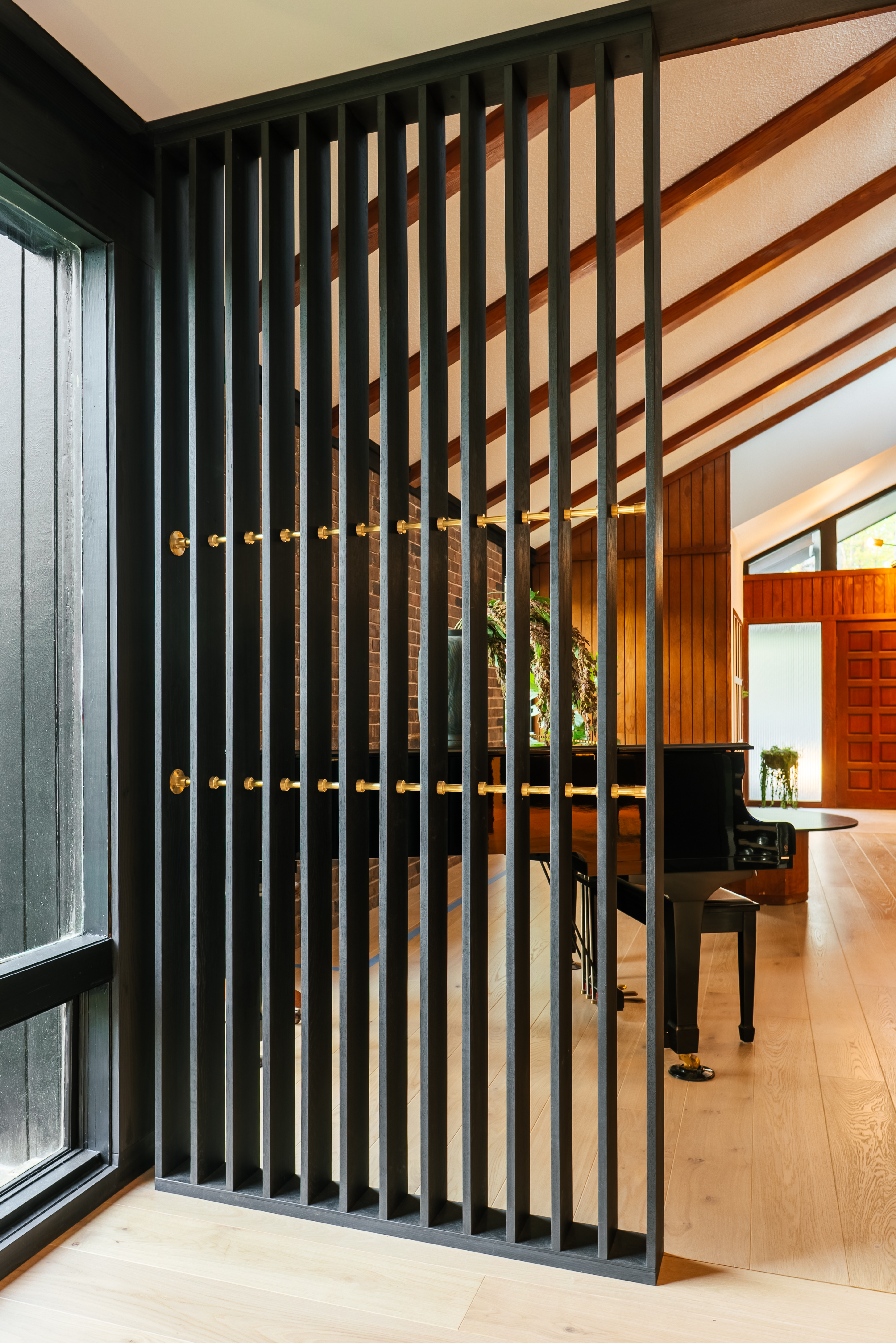 A black slatted room divider with brass details stands in the foreground, separating a light wood floor entryway from a living area featuring a black grand piano, a brick fireplace, and a vaulted ceiling with exposed wooden beams.
