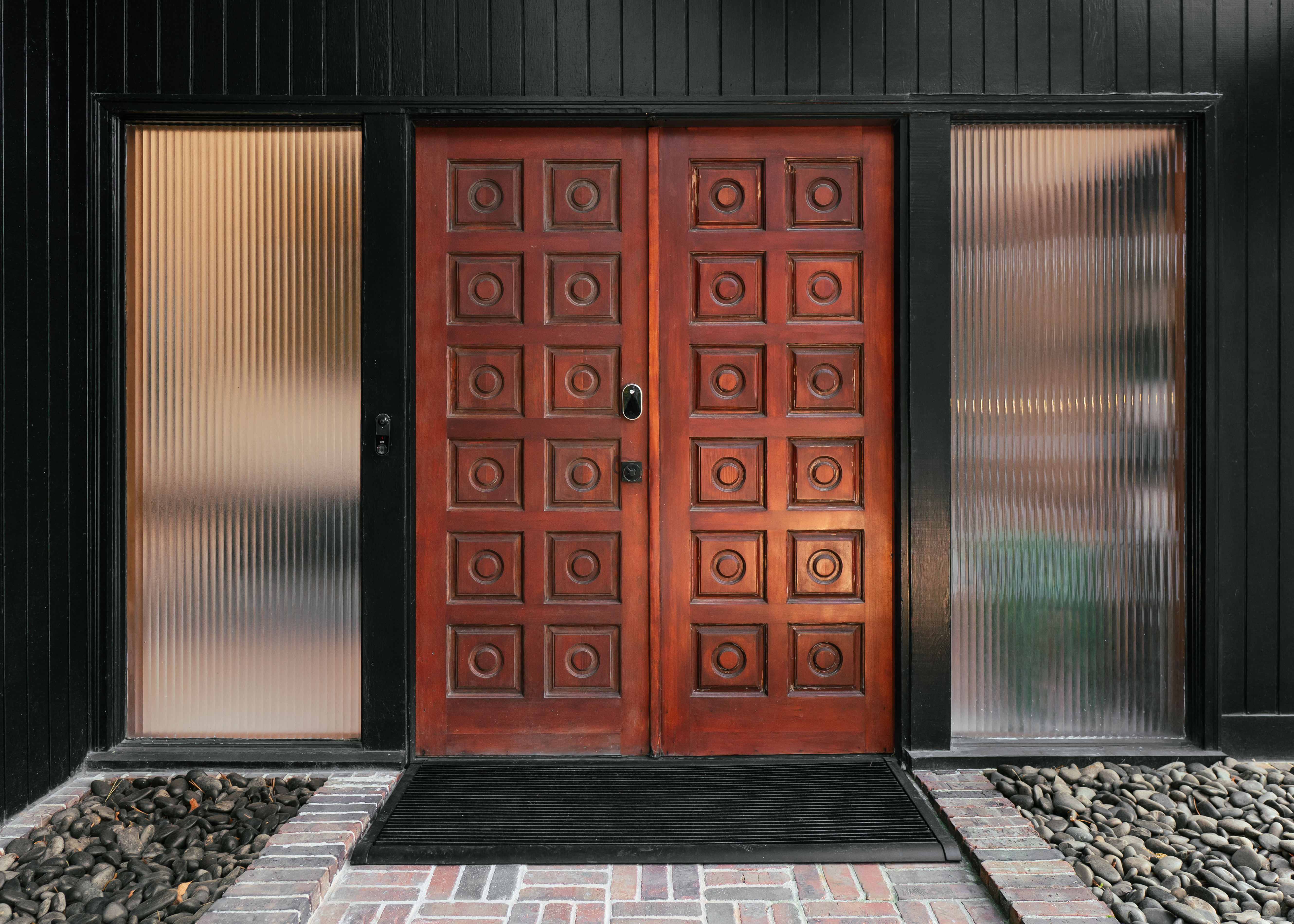 A close-up of the front entrance of Ravenwood, featuring dark wooden double doors with square and circular carved panels, flanked by tall vertical fluted glass sidelights, all set against a dark exterior wall with a brick and pebble walkway.