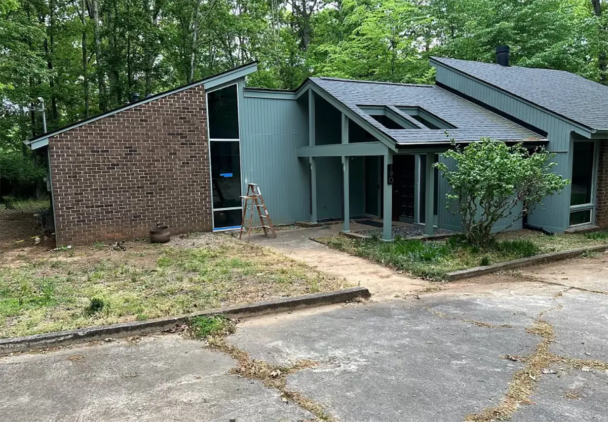 A mid-century modern home named Ravenwood in Athens, GA, featuring a distinctive angular design with dark brick and teal vertical siding, large floor-to-ceiling windows, and a prominent front entrance with skylights, surrounded by a natural, wooded landscape.