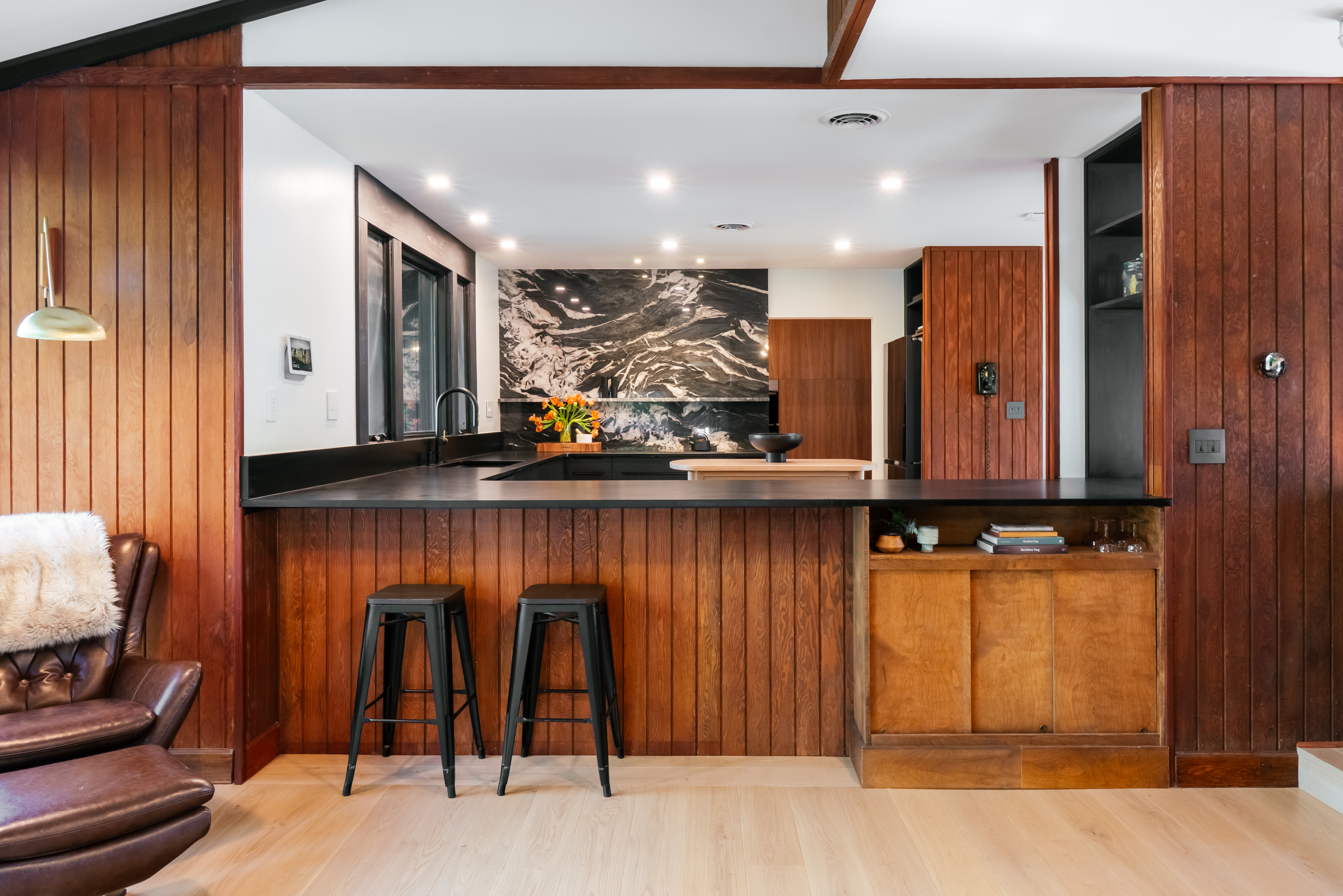 A mid-century modern kitchen in Athens, GA, featuring rich vertical wood paneling, black countertops, two black bar stools, and a dramatic black and white marble backsplash, illuminated by recessed lighting.