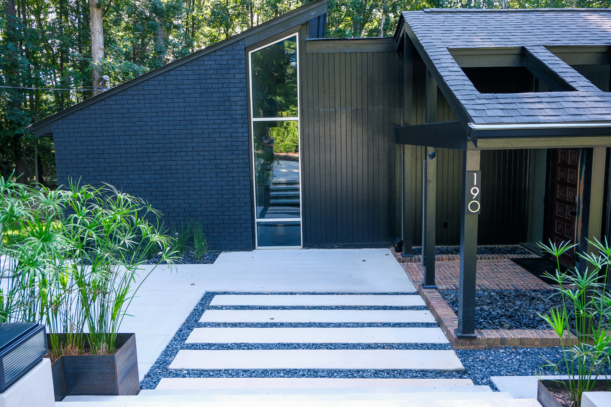 The front exterior of a dark, angular mid-century modern home named Ravenwood, featuring black brick and vertical siding, a tall vertical window, and a modern concrete pathway with dark grey gravel leading to the entrance.