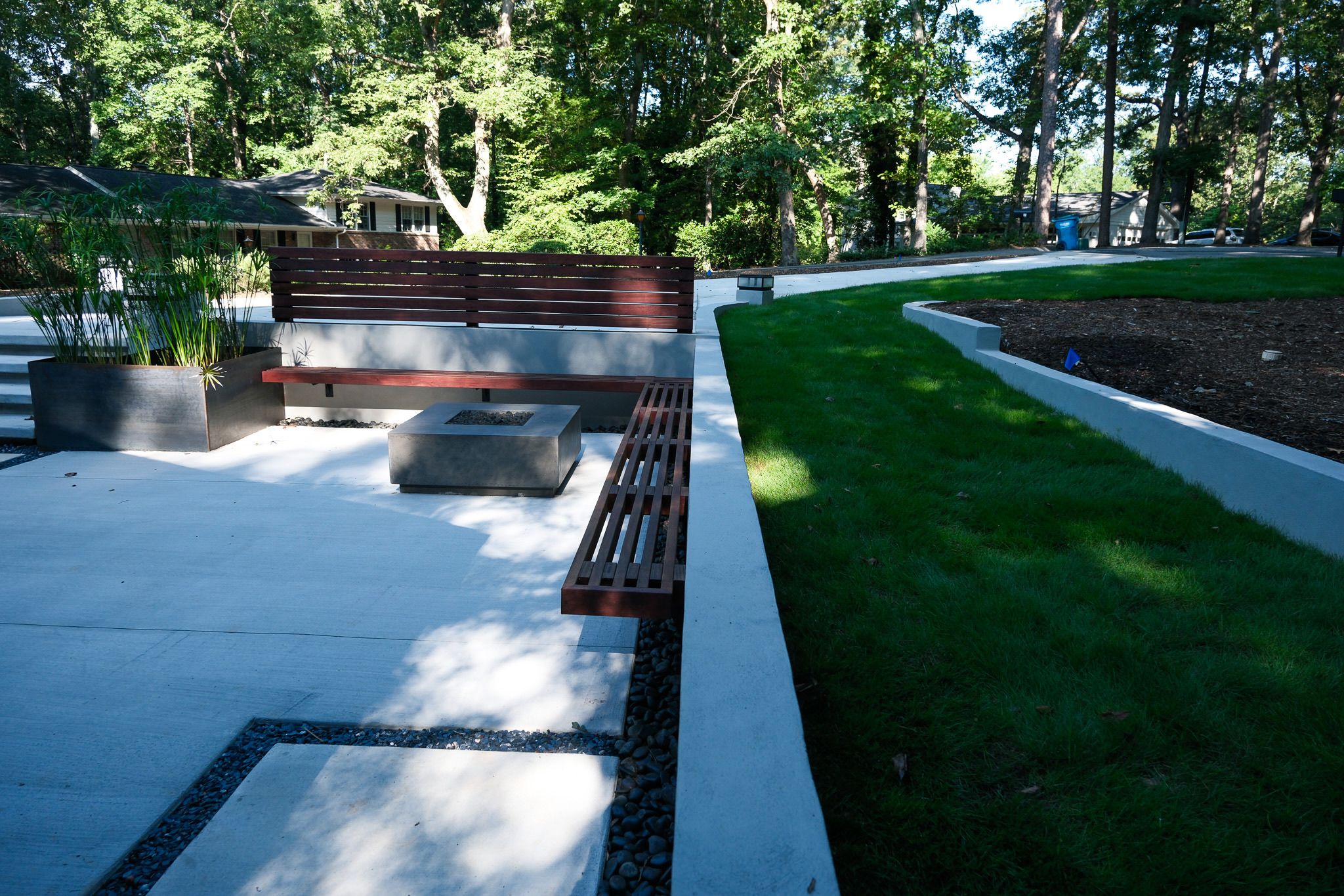 A modern outdoor living space at Ravenwood features a light gray concrete patio with integrated dark wood slatted benches, a concrete fire pit, and a large dark metal planter with tall green plants, bordered by a manicured lawn and mature trees.