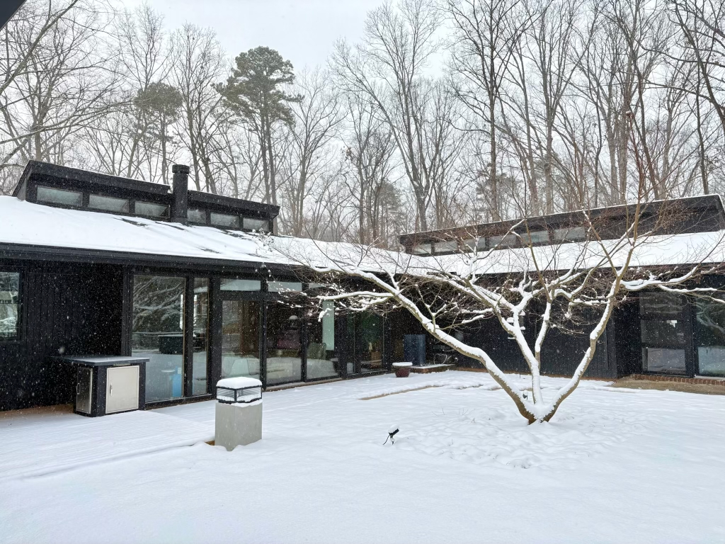 A mid-century modern home named Ravenwood, with a dark exterior and large windows, is covered in a fresh blanket of snow, surrounded by bare trees under a grey winter sky.