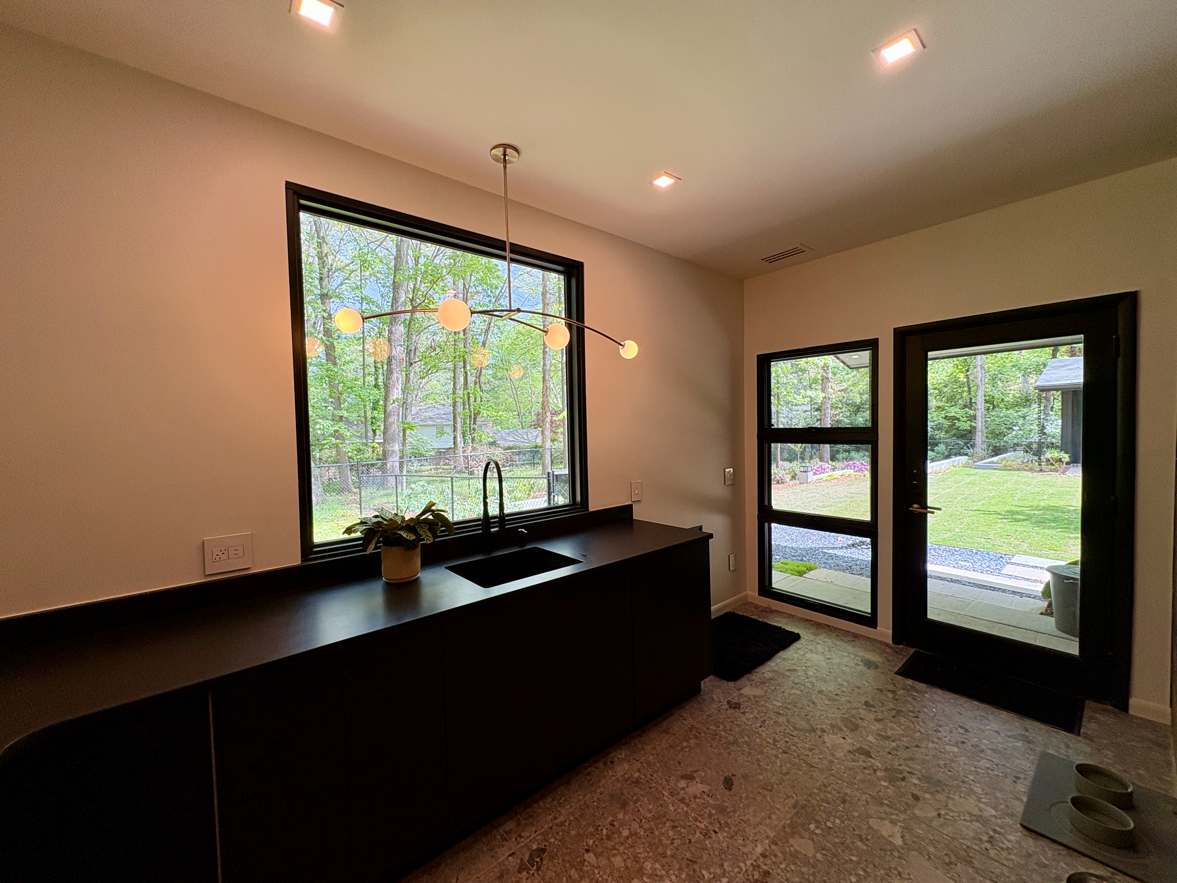 A modern utility room features dark cabinetry, a black sink, and a stylish chandelier, with large windows and a glass door offering views of a vibrant green backyard and surrounding trees.
