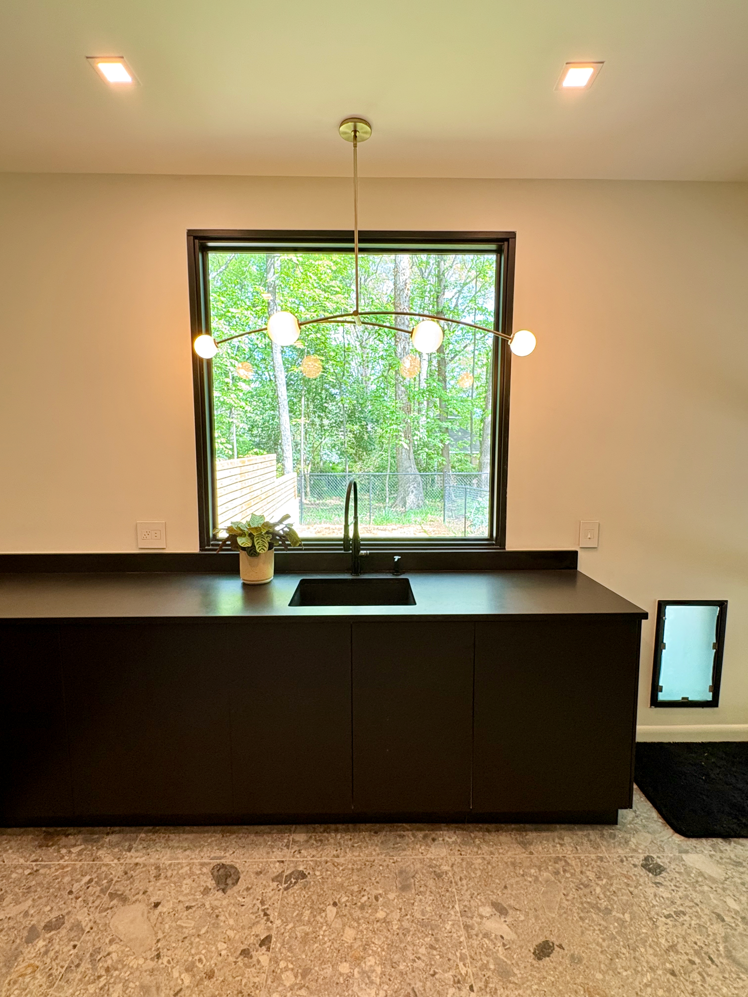 A modern kitchen area features dark, flat-panel cabinetry with an integrated black sink and faucet, beneath a large window showcasing vibrant green trees. A contemporary brass chandelier with spherical lights hangs above the sink, and light-colored terrazzo flooring extends across the space.