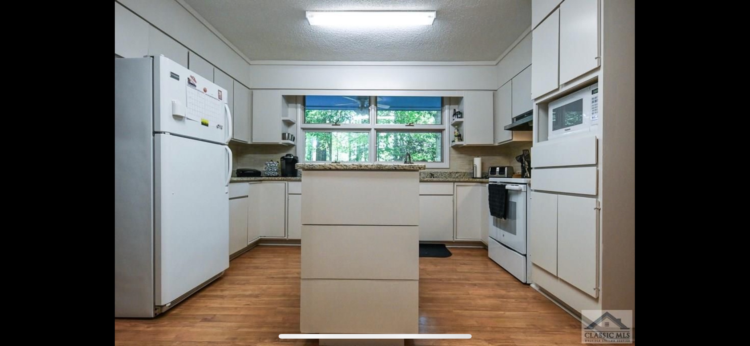A functional kitchen with white cabinets and appliances, wood-look flooring, and a central island, featuring a large window overlooking lush trees.