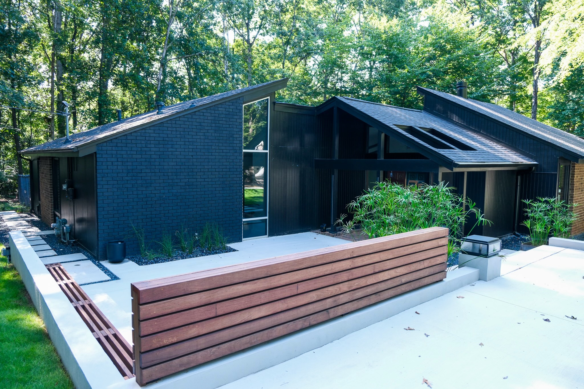 A mid-century modern home named Ravenwood, featuring a dark blue brick and black vertical siding exterior with angular rooflines and large windows, is set against a backdrop of dense green trees, with a modern wooden slatted fence and concrete pathway in the foreground.
