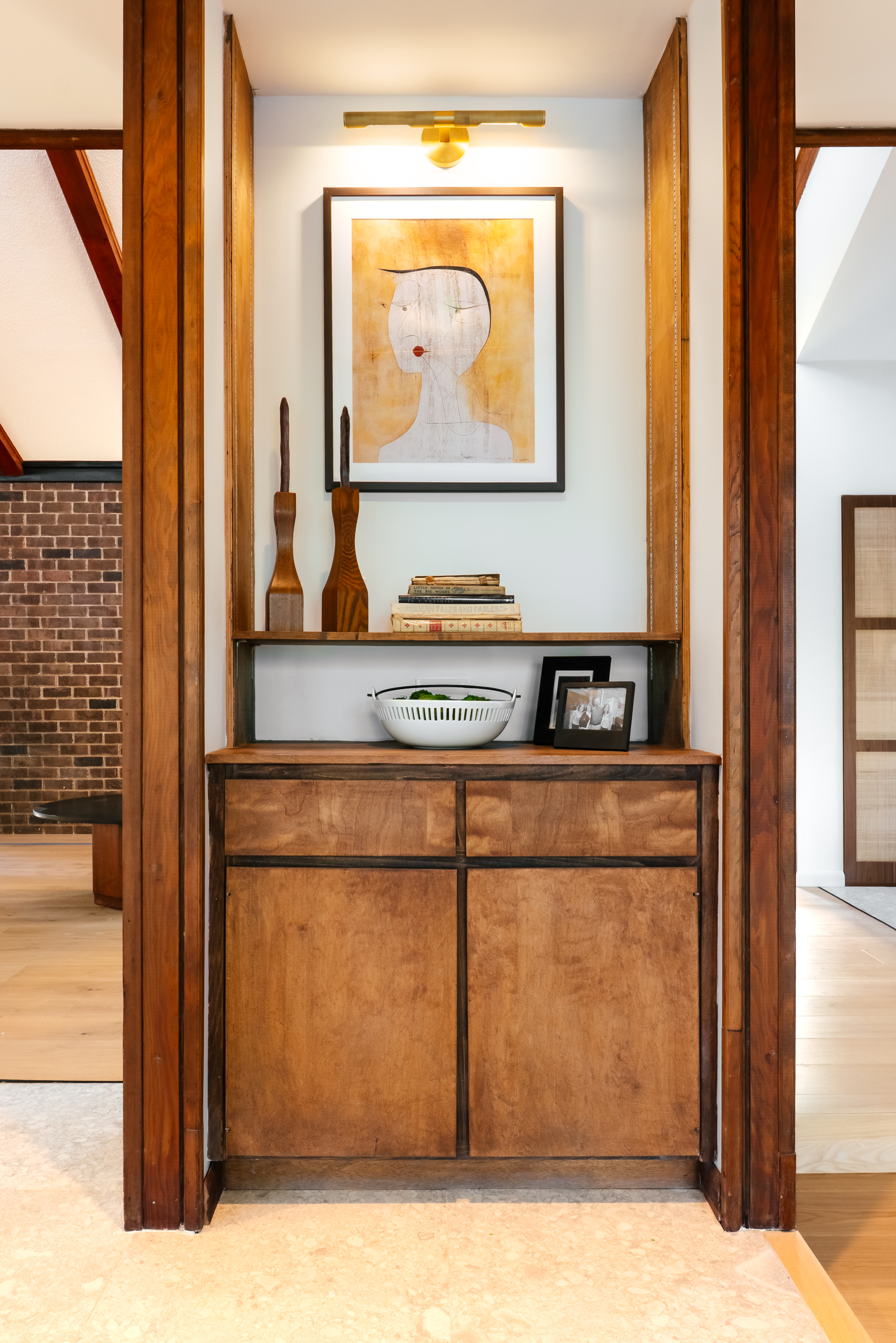 A warm, built-in wooden cabinet with shelves displays an abstract framed artwork of a woman's face, wooden sculptures, books, and framed photos, illuminated by an overhead brass light fixture.