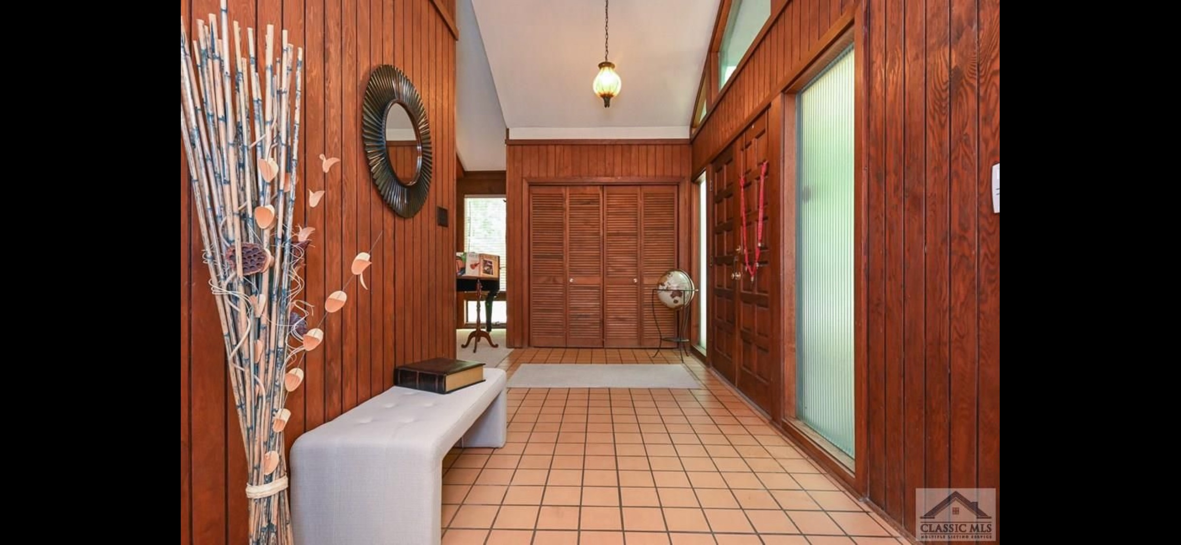 A mid-century modern entryway features extensive reddish-brown wood paneling on the walls, a terracotta tiled floor, and a light gray upholstered bench. A round mirror hangs on the left wall, and a large wooden front door with a side panel of fluted glass is visible on the right, with louvered bi-fold doors at the far end of the hall.