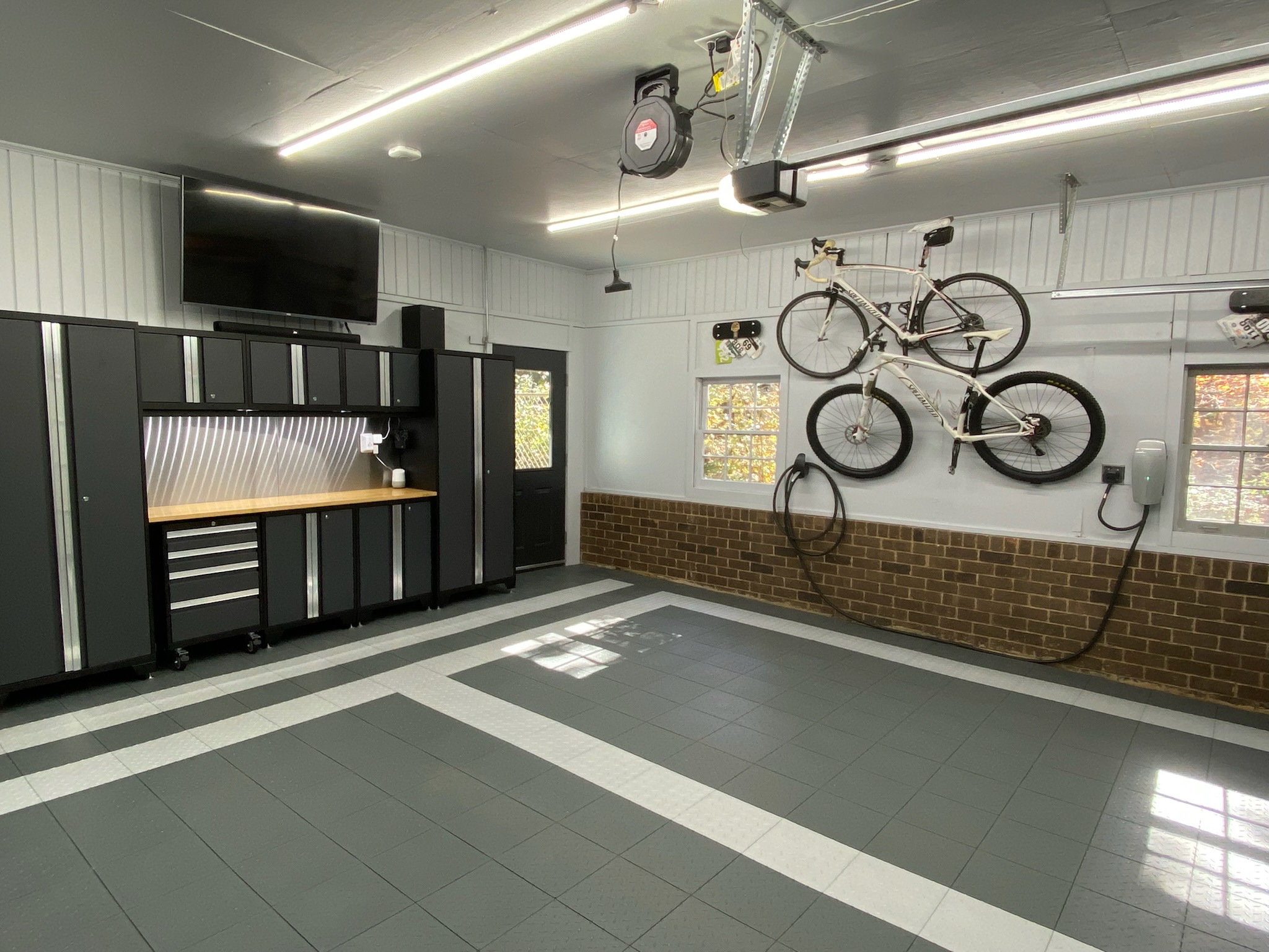 A meticulously organized modern garage with dark gray modular cabinets, a light wood workbench, gray and white checkered floor tiles, two bicycles hanging on the wall, and windows with brick detailing.