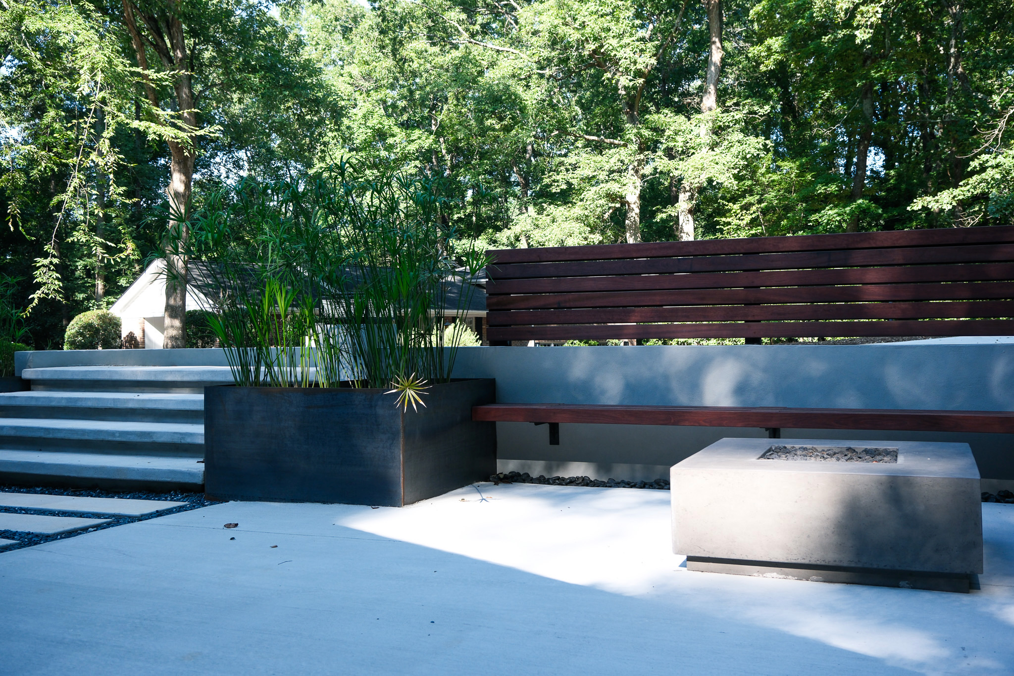 A modern outdoor patio with concrete steps, a dark metal planter overflowing with tall green reeds, a wooden slatted bench integrated into a low concrete wall, and a rectangular concrete fire pit, all set against a backdrop of lush green trees and dappled sunlight.