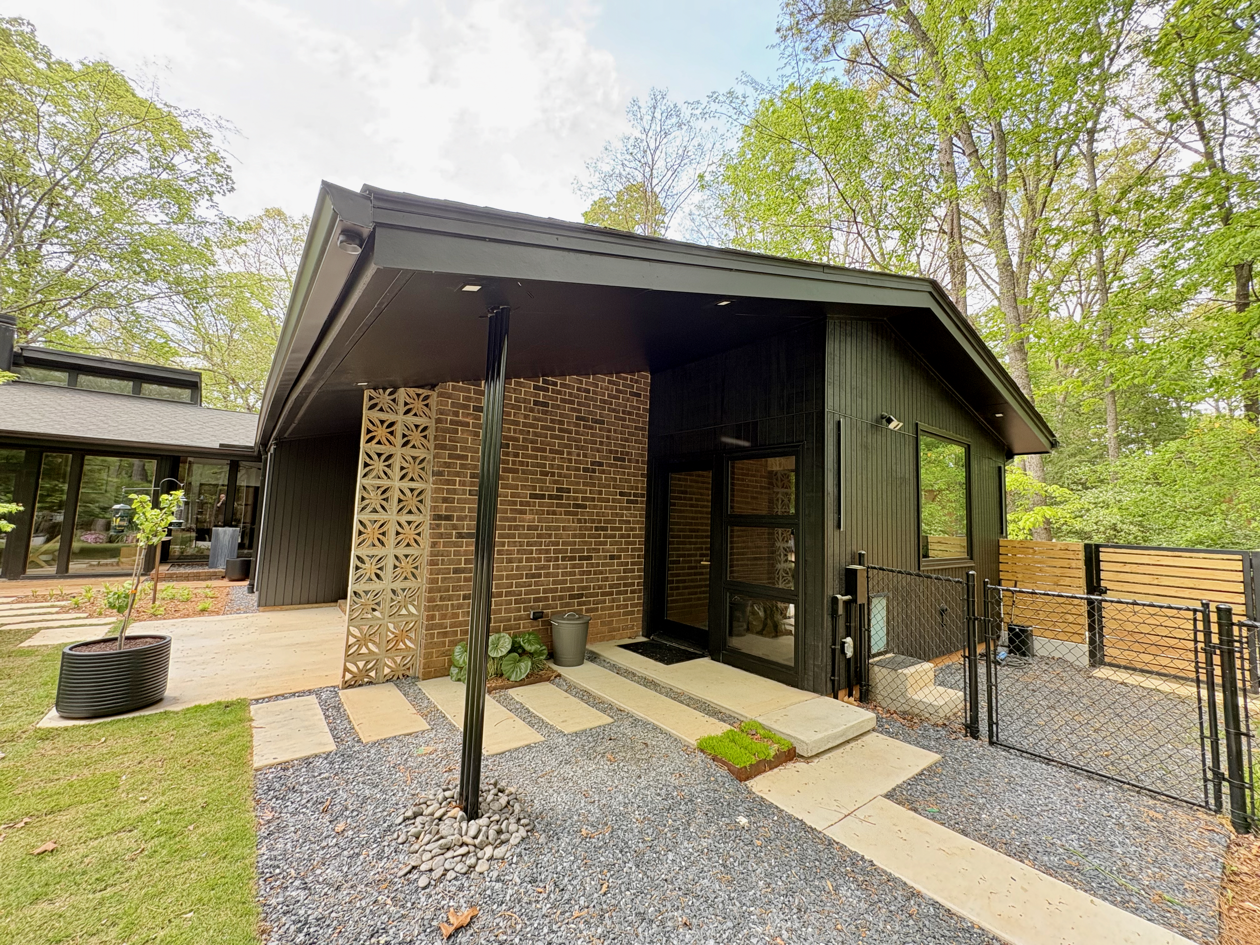 Exterior view of Ravenwood, a mid-century modern home in Athens, GA, featuring a dark wood facade, exposed brick, and decorative breeze blocks, with a concrete and gravel walkway leading to the glass-paneled front door, all set amidst vibrant green trees and landscaping.