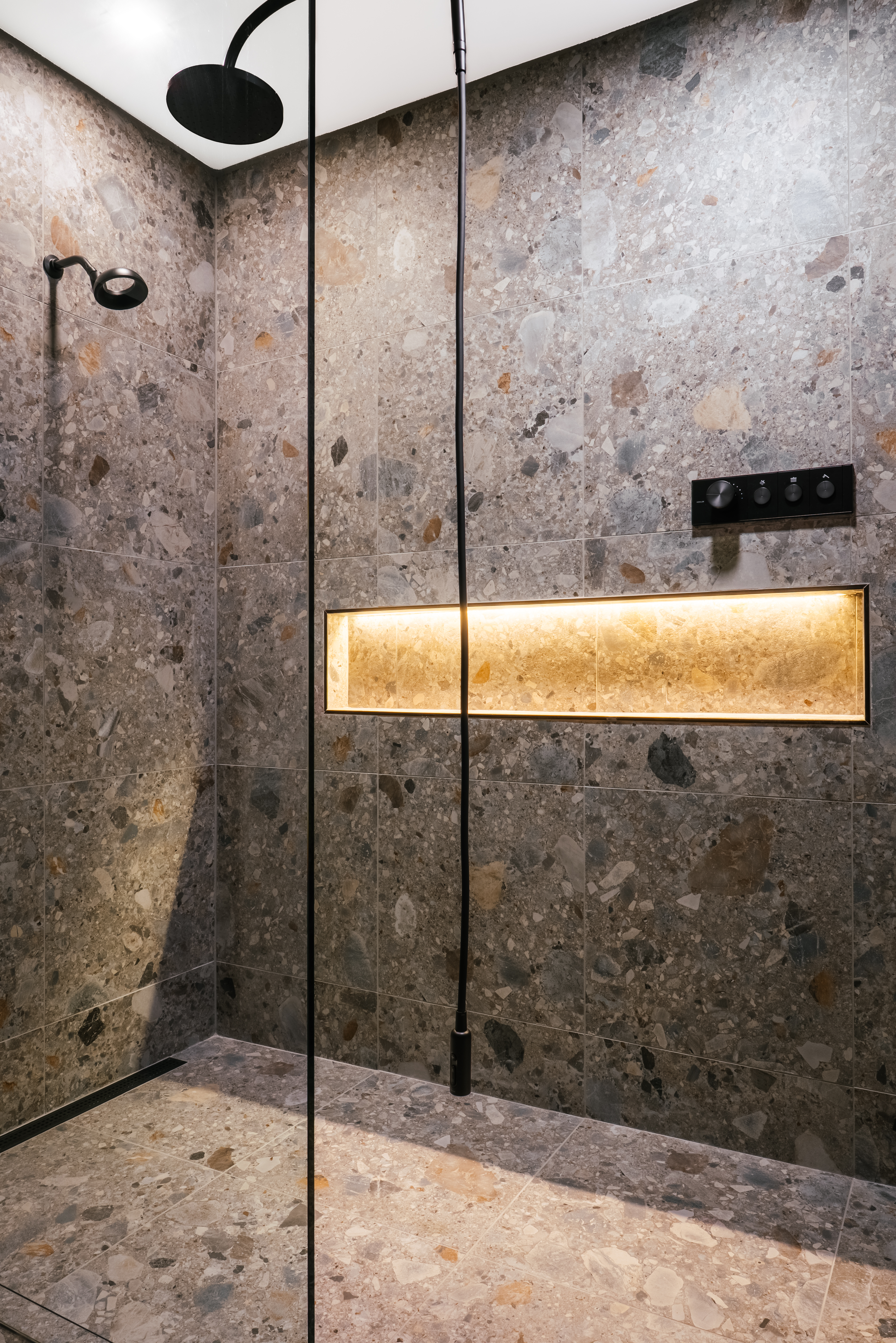 A luxurious modern shower with grey speckled terrazzo-style tiles covering the walls and floor, featuring matte black rain and handheld showerheads, black digital controls, and a long, glowing amber-lit niche.