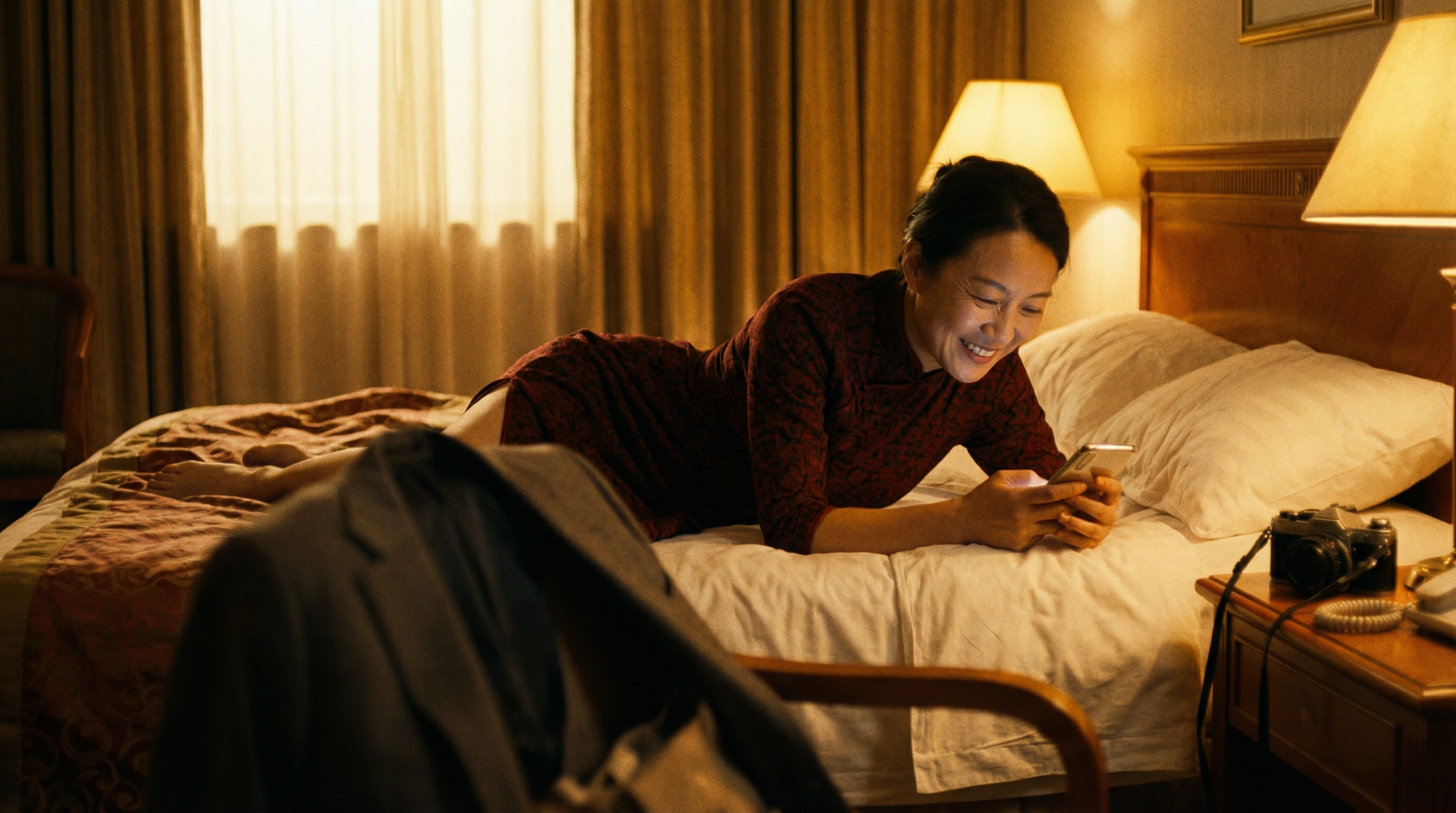 A woman lying on a hotel bed, smiling at a message on her phone