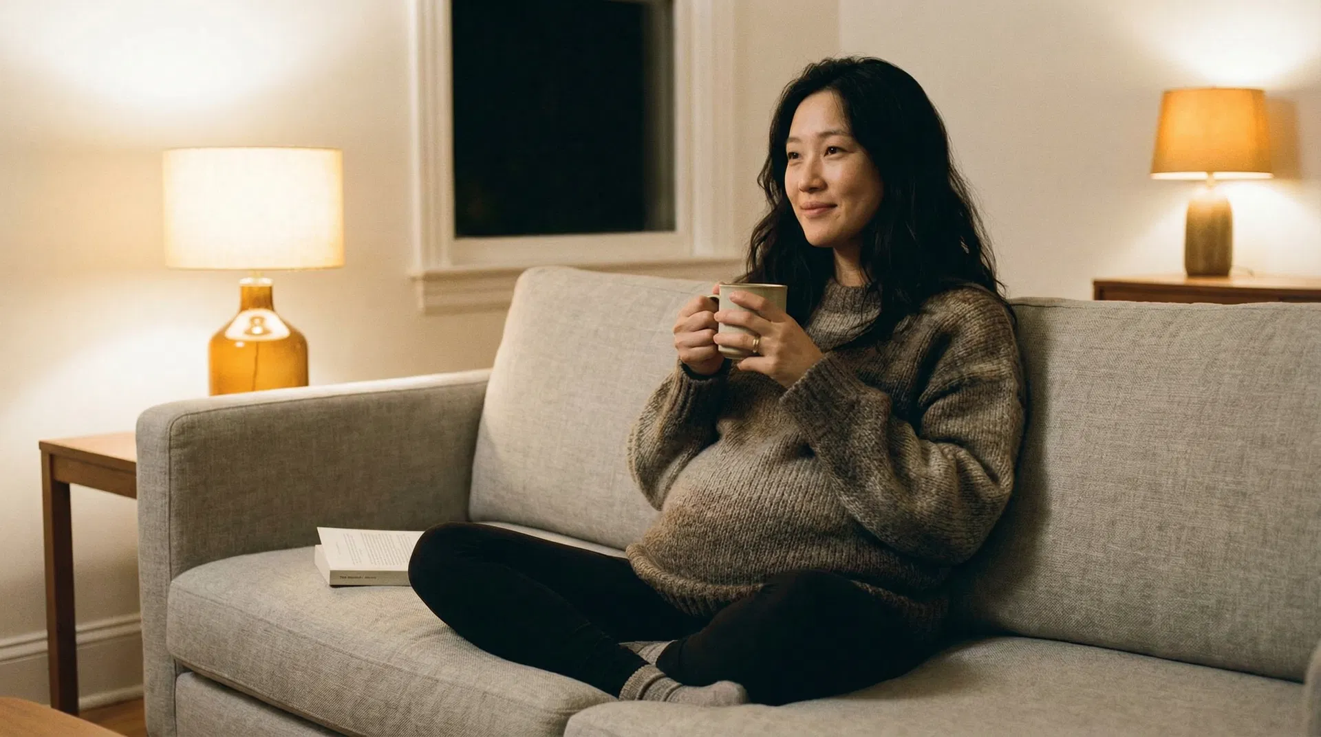 Pregnant Asian woman curled up in armchair with tea, quietly contemplating