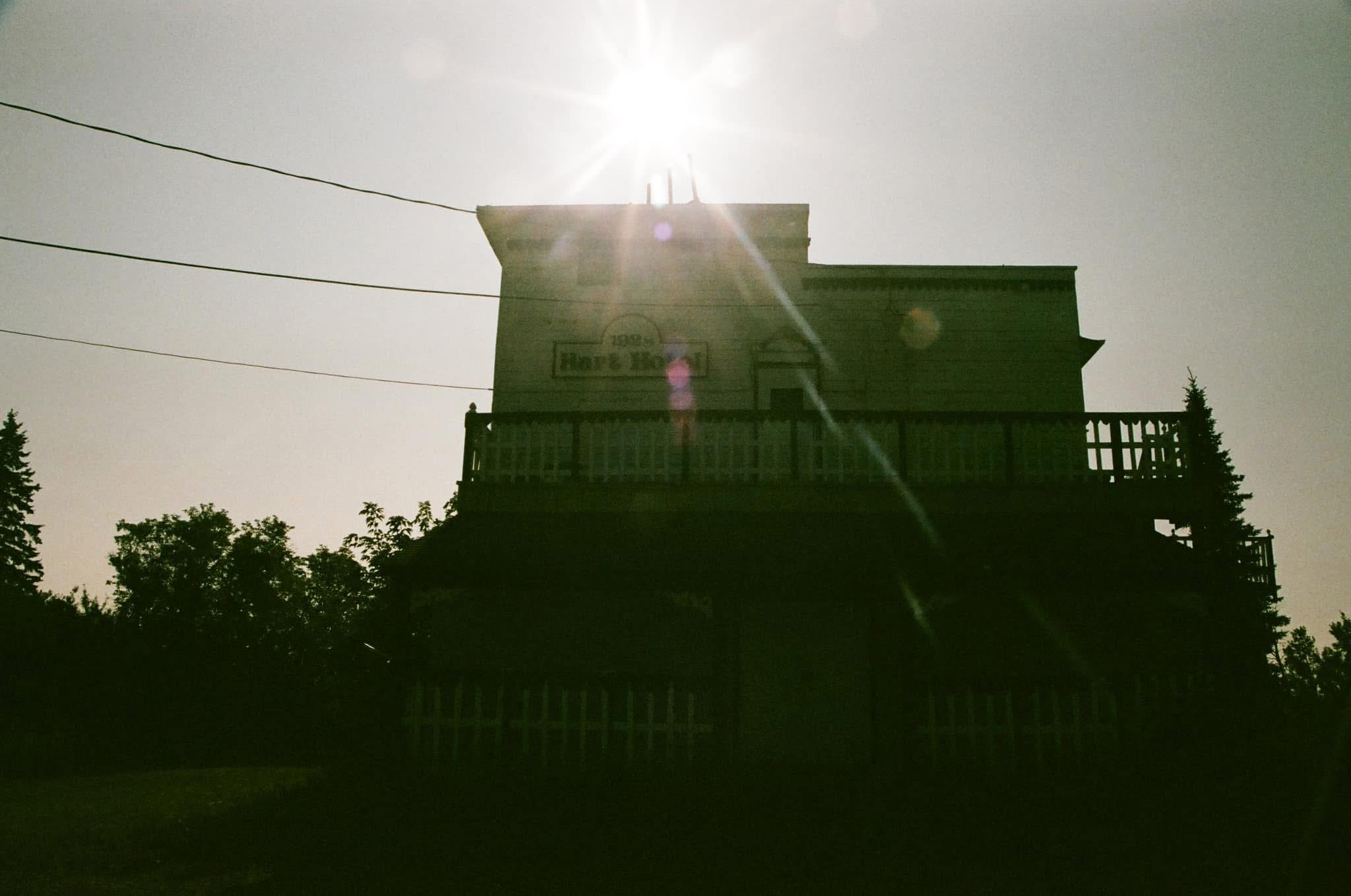 analog film photograph of an old wooden building silhouetted against a blazing sun, power lines cutting across the sky