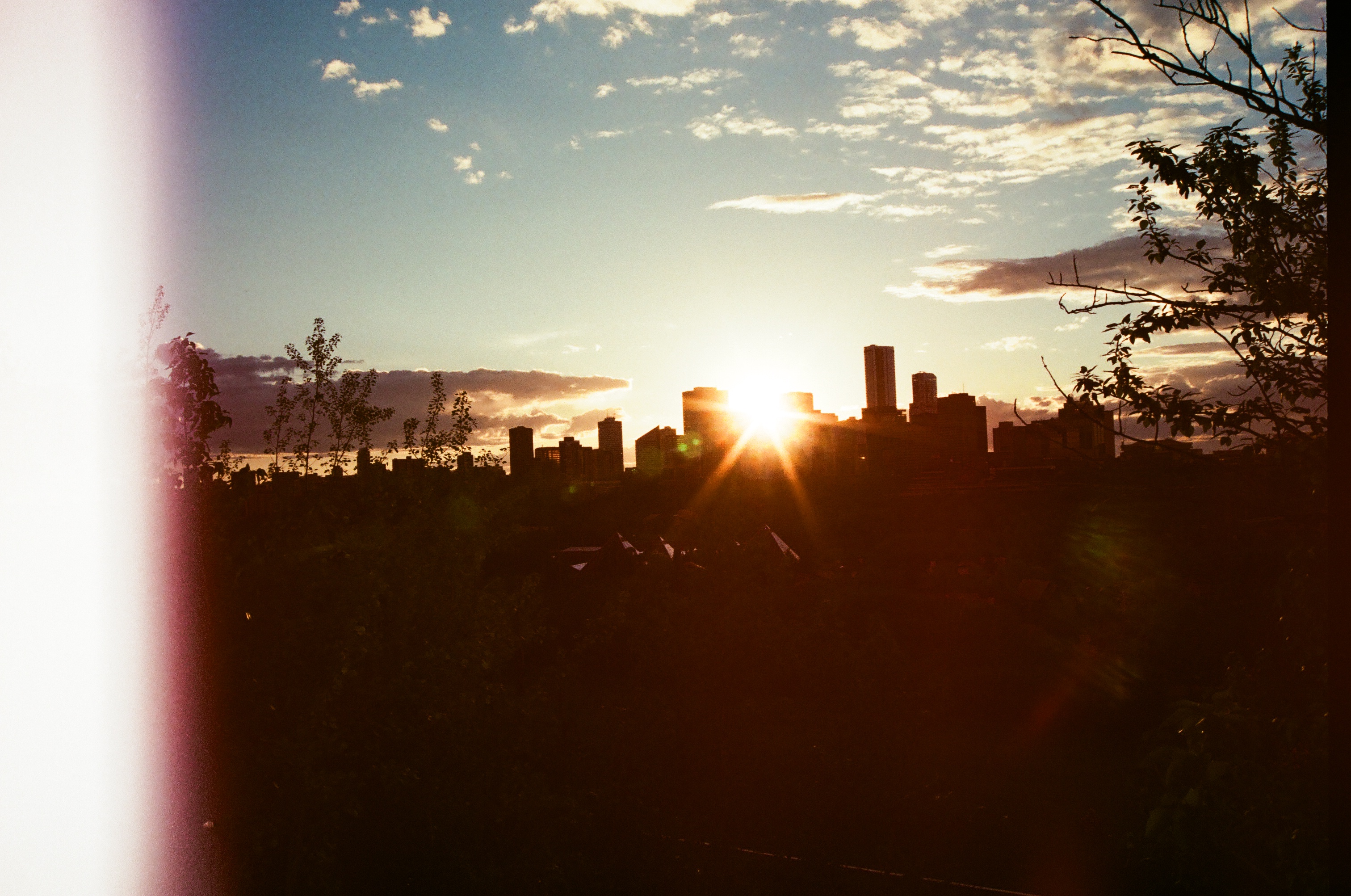 analog film photograph of a city skyline silhouetted against a golden sunset, with lens flare and light leak on the left edge