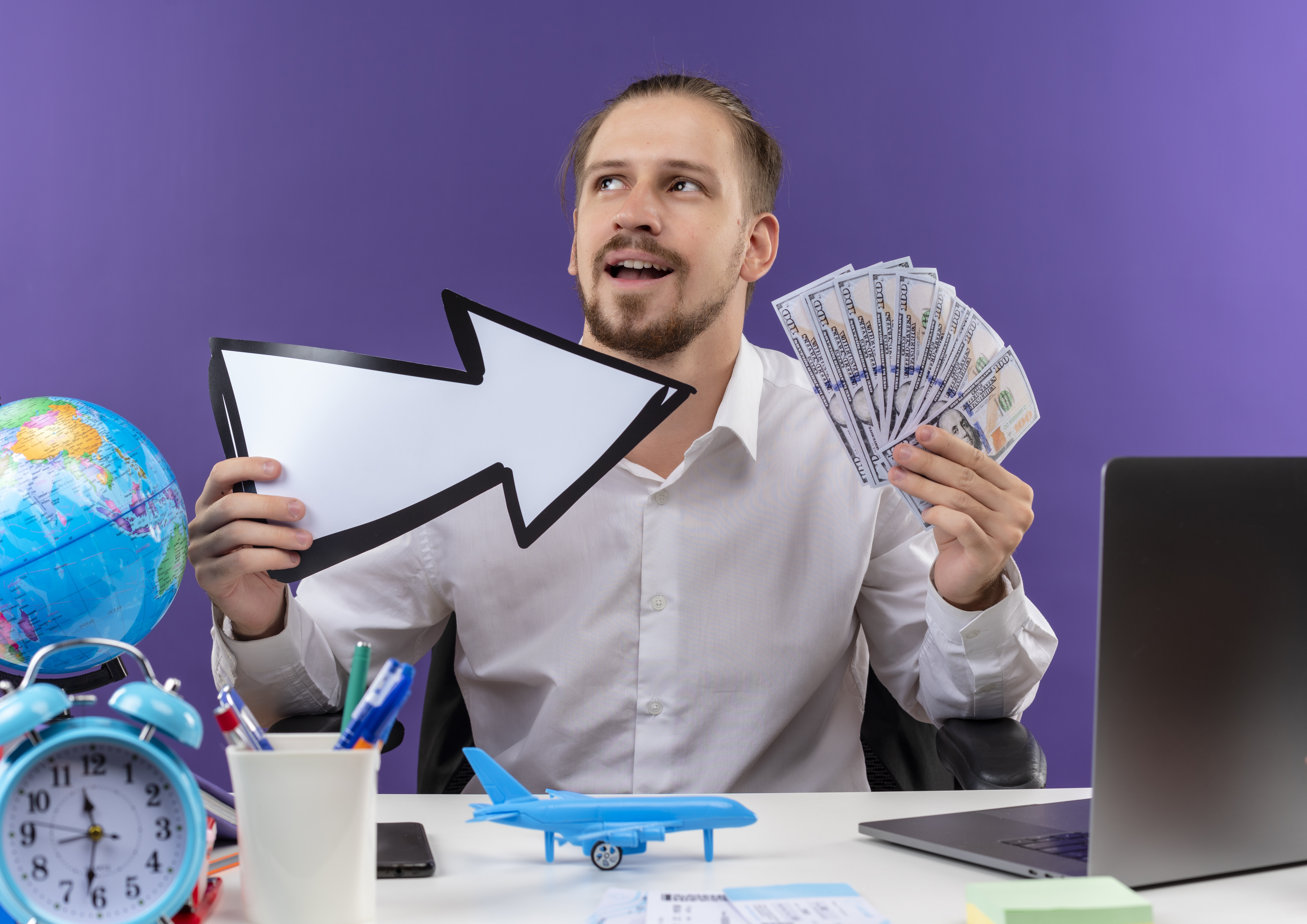 Handsome businessman in white shirt holding white arrow showing cash looking aside with smile on face sitting at the table in offise over purple background