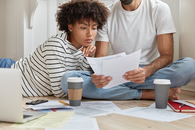 Hard working interracial marketing experts pose on floor, study documentation, make monthly report
