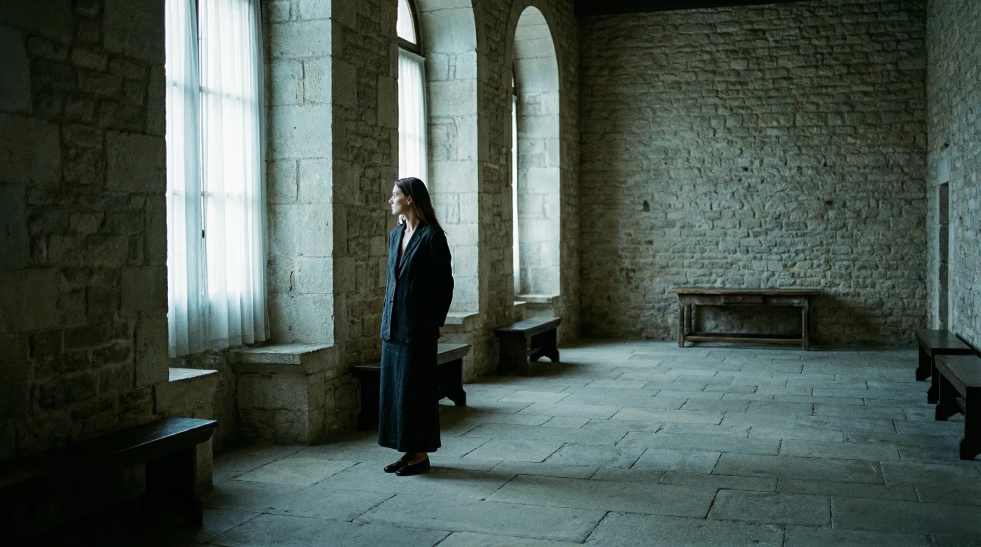 A woman standing alone in a stone-walled hall with arched windows, facing the light — the stillness before a decisive response
