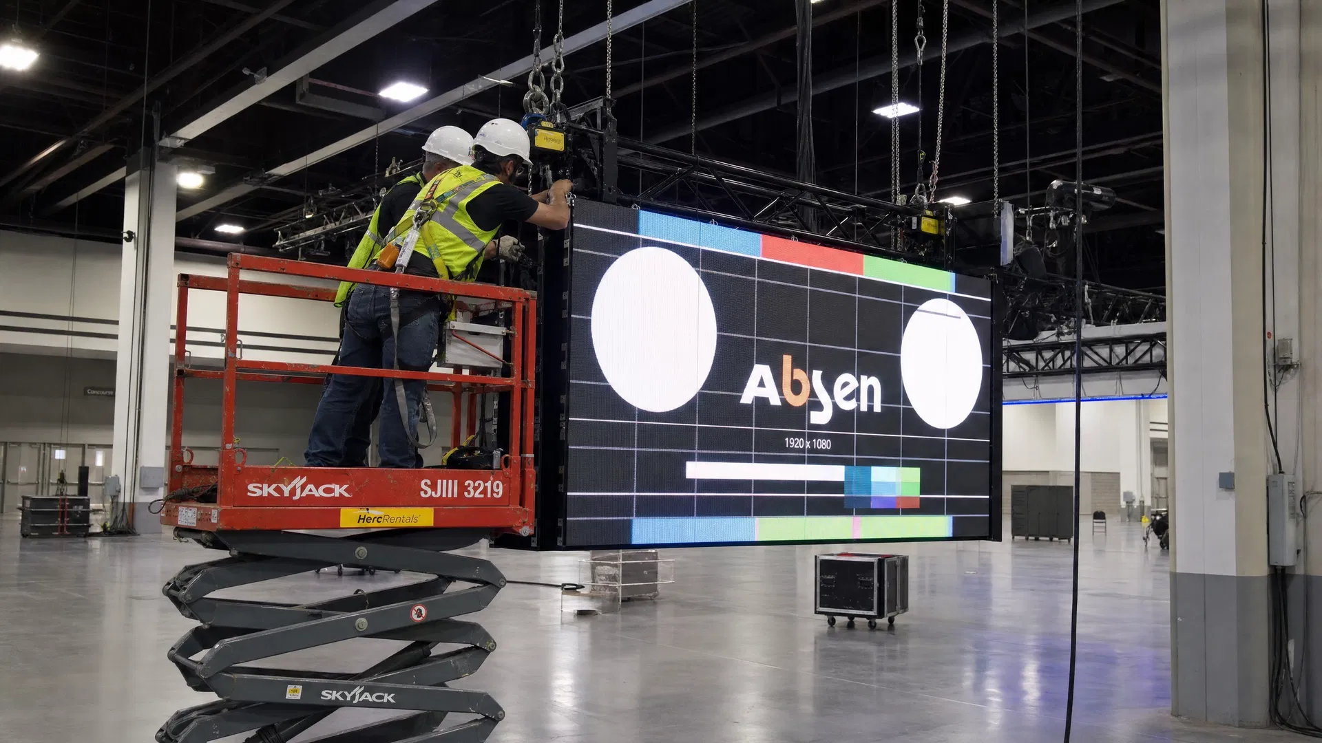 Professional riggers installing an LED hanging sign at a trade show convention center using a scissor lift and chain motors