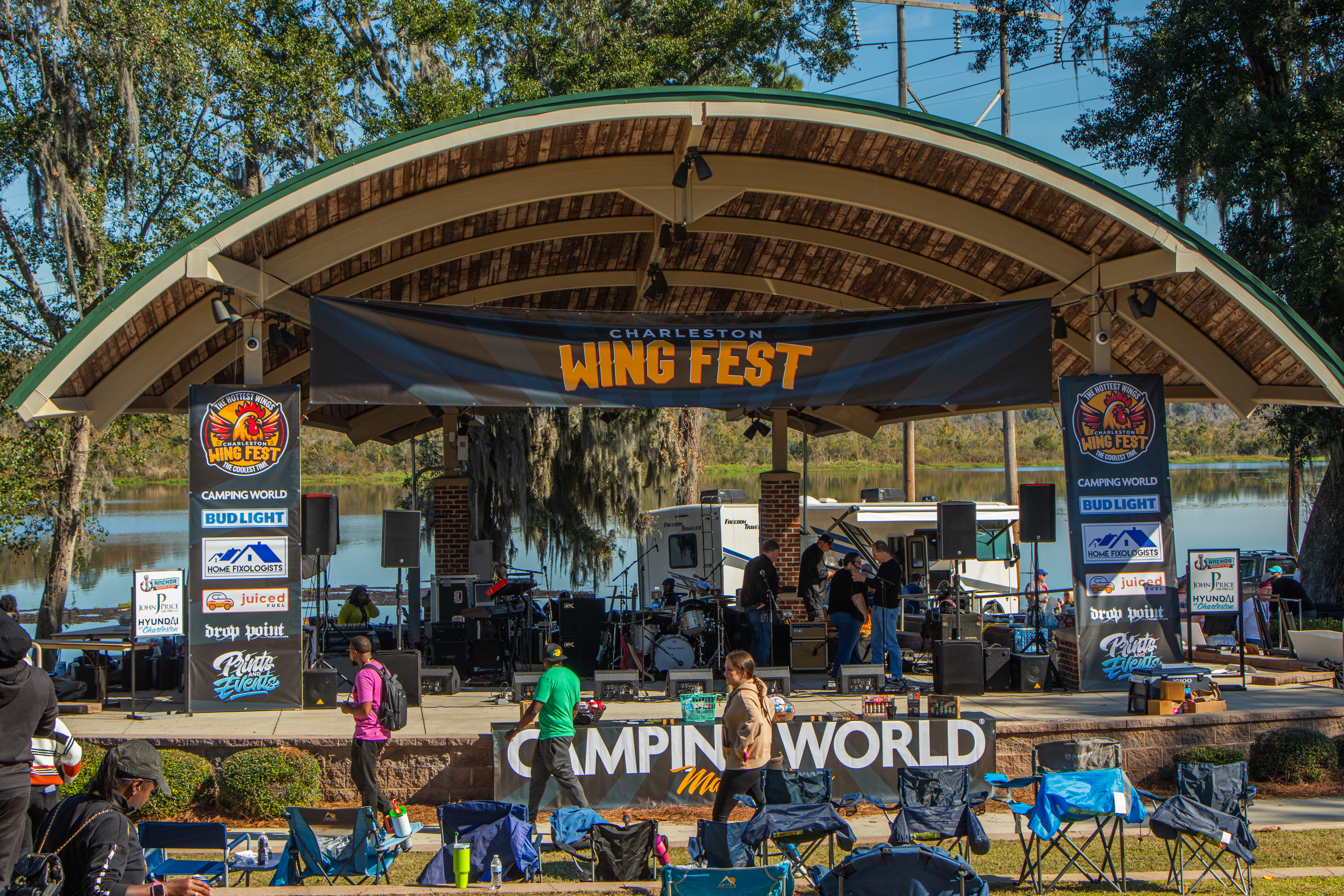 Charleston Wing Fest stage with sponsor banners, musicians, and attendees by a lake.