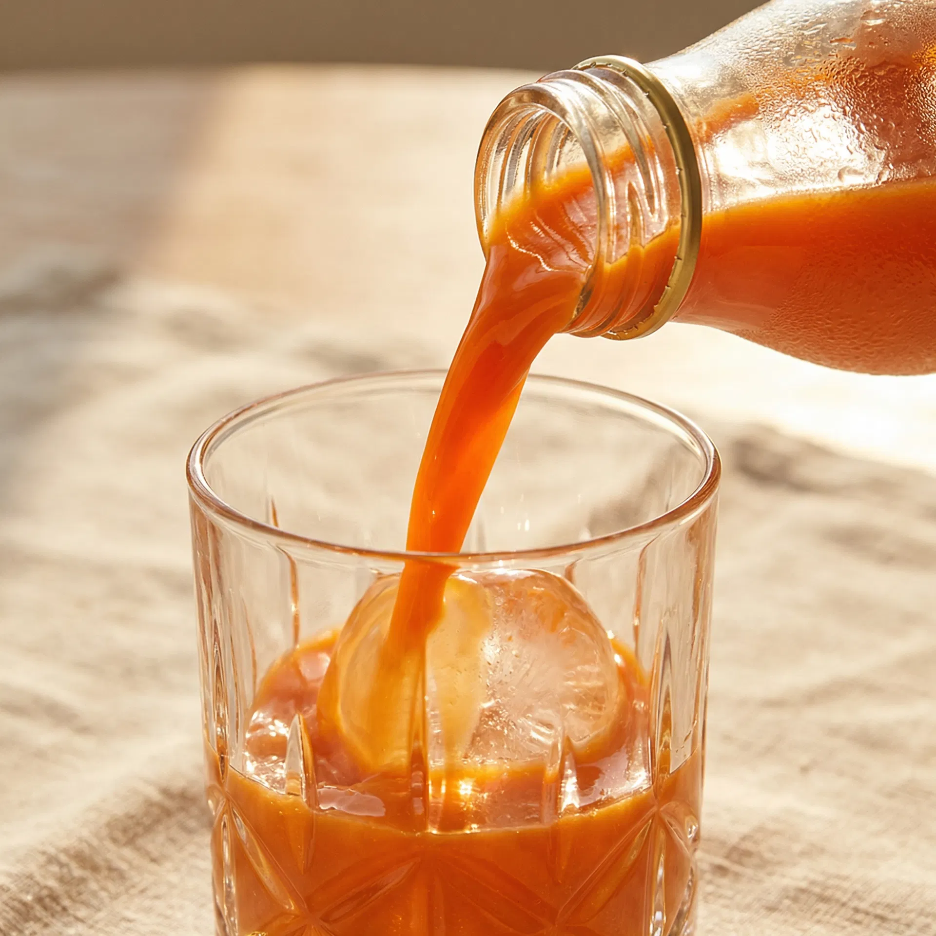 Orange carrot juice being poured from a gold-lid bottle into a crystal glass over ice