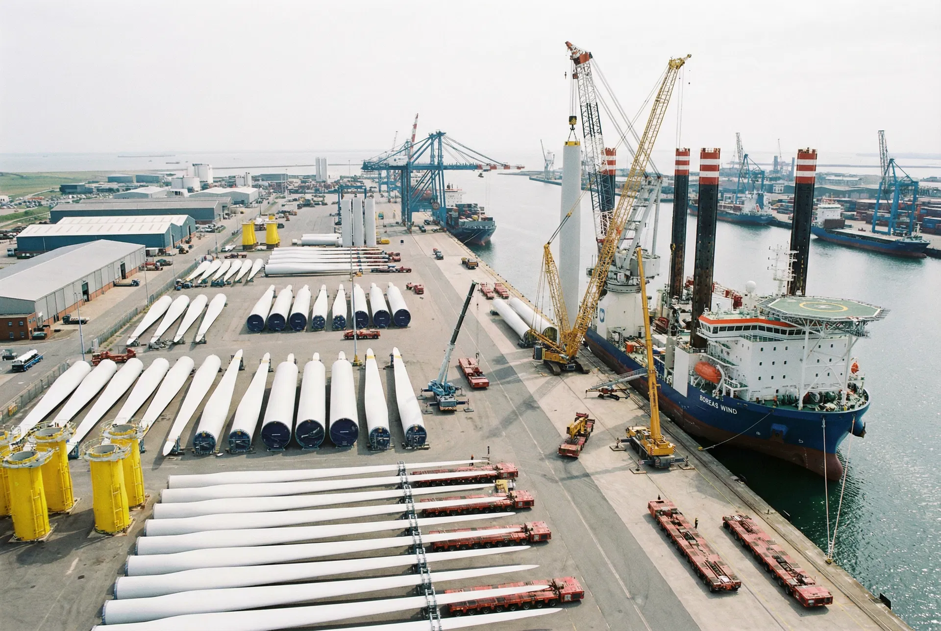 Aerial view of offshore wind staging port with turbine blades, towers, and installation vessel