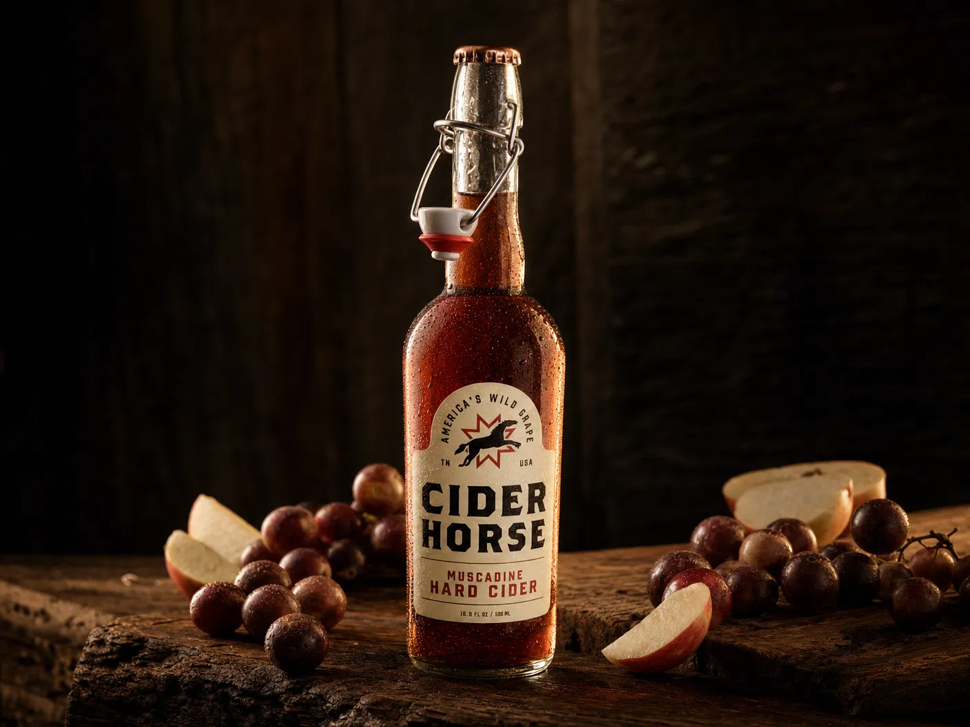 Cider Horse Red Muscadine Hard Cider swing-top bottle with condensation on a dark wood surface with muscadine grapes and apple slices