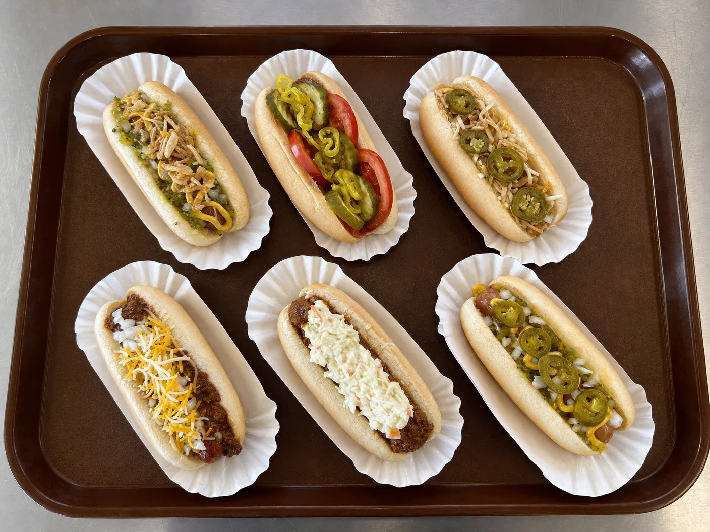 Overhead lineup of six Franken Dogs hotdogs with chili, cheese, slaw, peppers, pickles, tomatoes, and onions