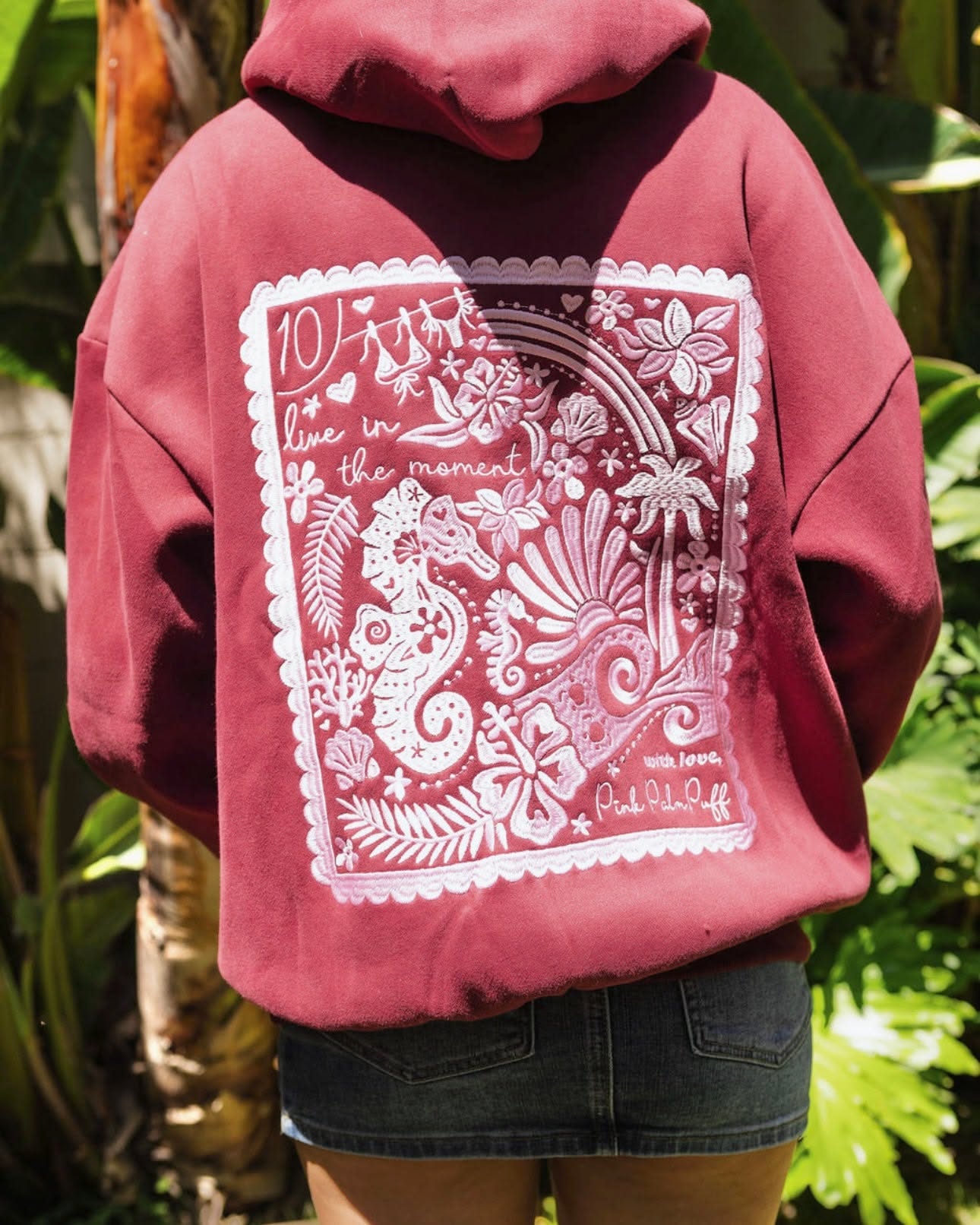 Pink Palm Puff maroon red hoodie — pullover style, drawstring hood, large white and pink graphic print on the back featuring a seahorse, tropical flowers, palm trees, and text 'live in the moment' and 'Pink Palm Puf