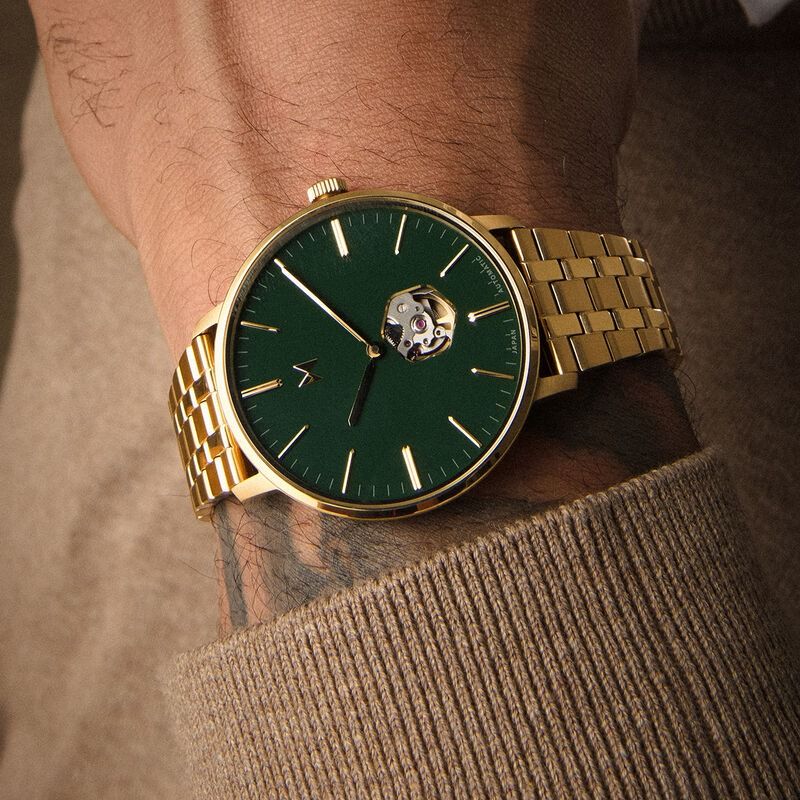 MVMT emerald green and gold-tone watch — round case, emerald green dial, gold-tone hour markers and hands, open heart dial showcasing automatic movement, gold-tone link bracelet, 'V' logo at 9 o'clock