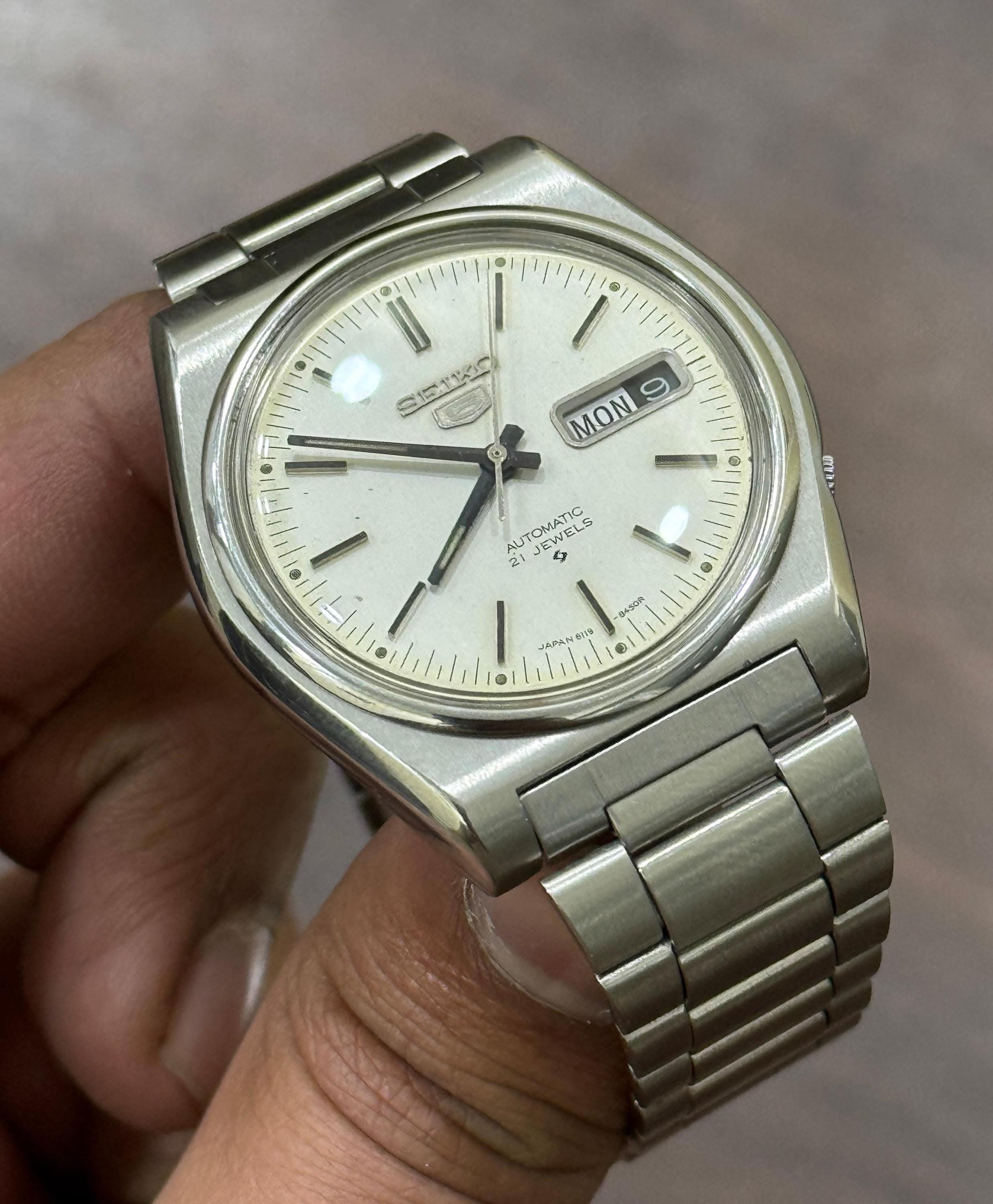 Seiko silver and off-white watch — day-date display at 3 o'clock, rectangular hour markers, dauphine hands, integrated bracelet, cushion-shaped case, automatic movement