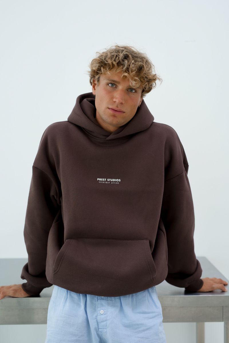 PREST STUDIOS dark brown hoodie — drawstring-free hood, kangaroo pocket, ribbed cuffs and hem, 'PREST STUDIOS EVERYDAY ATTIRE' text print on chest
